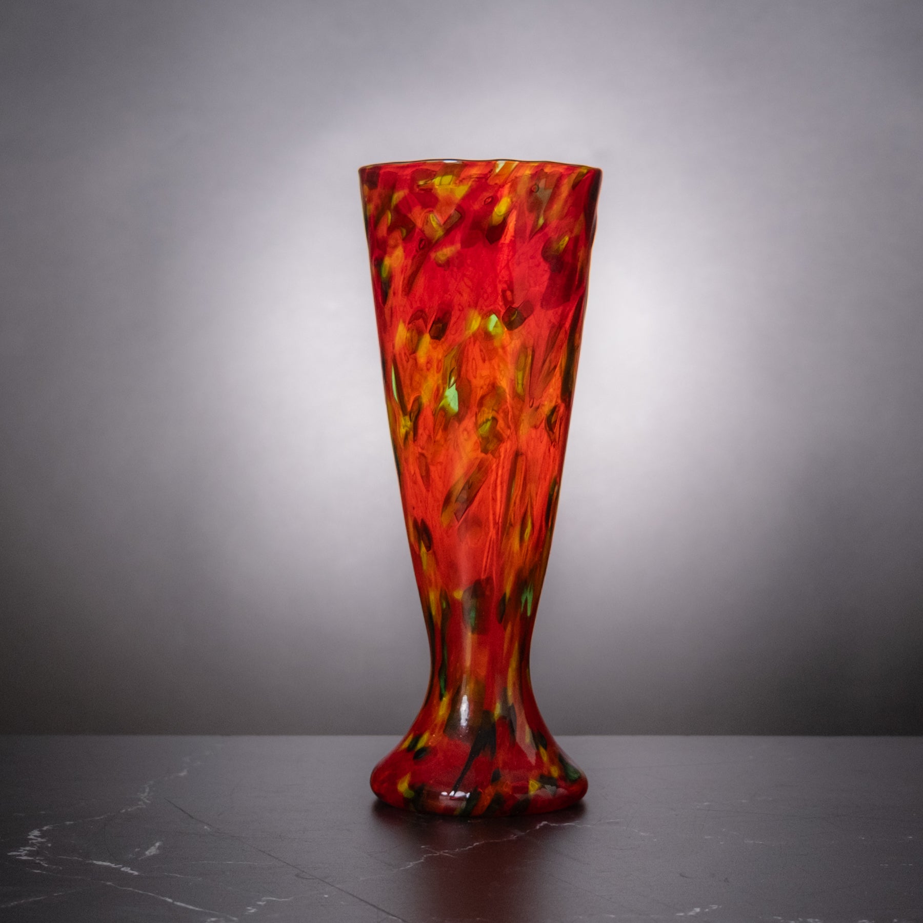 A tall, flared glass vase with a vibrant red, orange, and yellow marbled pattern, standing on a dark gray marble surface against a gray gradient background.