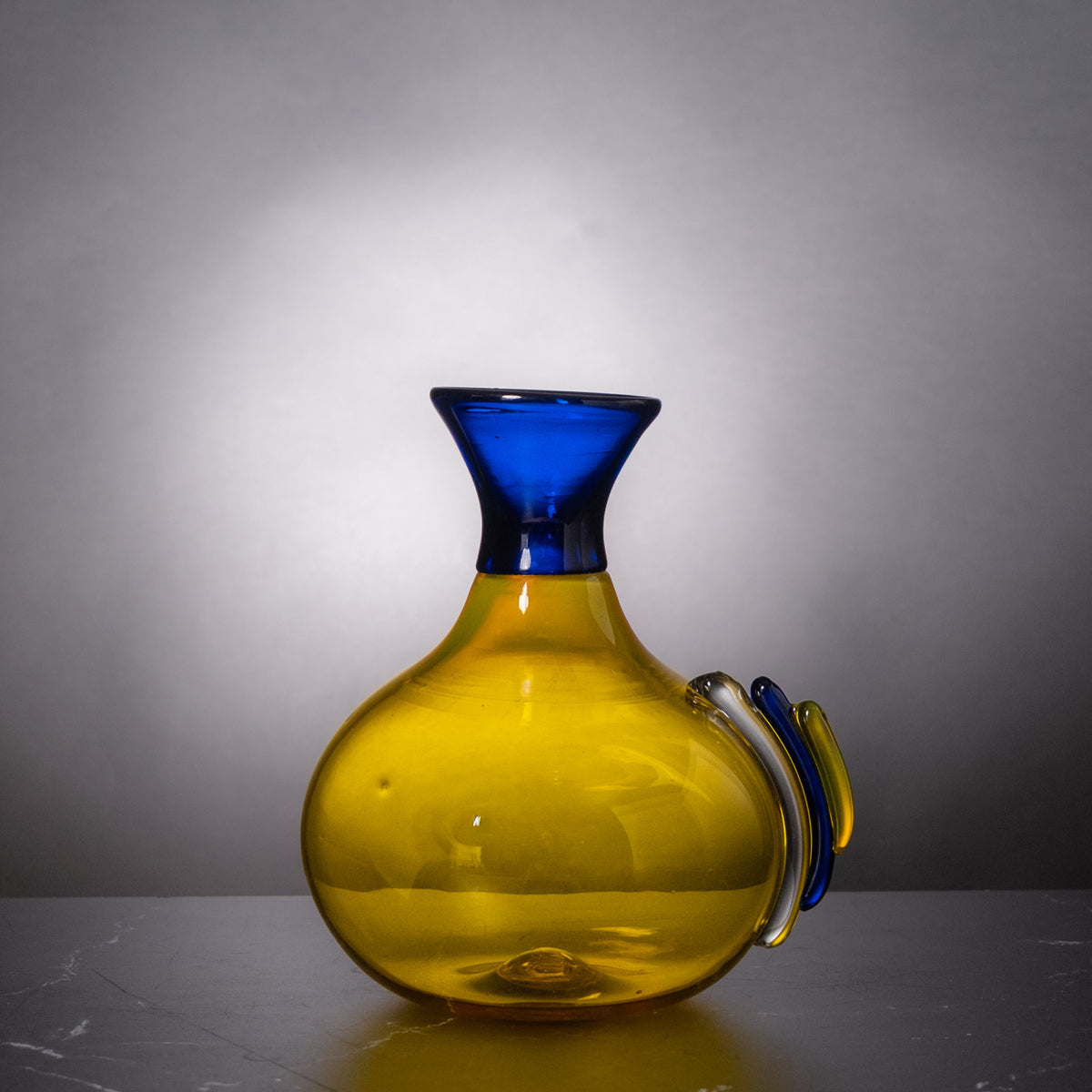 A hand-blown glass carafe with a bulbous yellow body, a flared cobalt blue neck, and two decorative glass handles in blue and white, set against a gray gradient studio background.