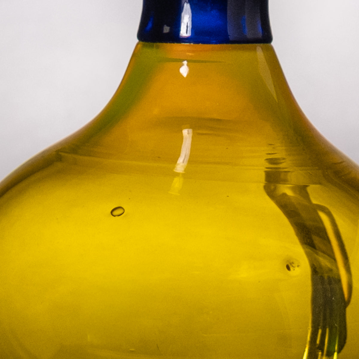 Close-up of a translucent yellow glass bottle or vase with a dark blue neck against a light gray background.