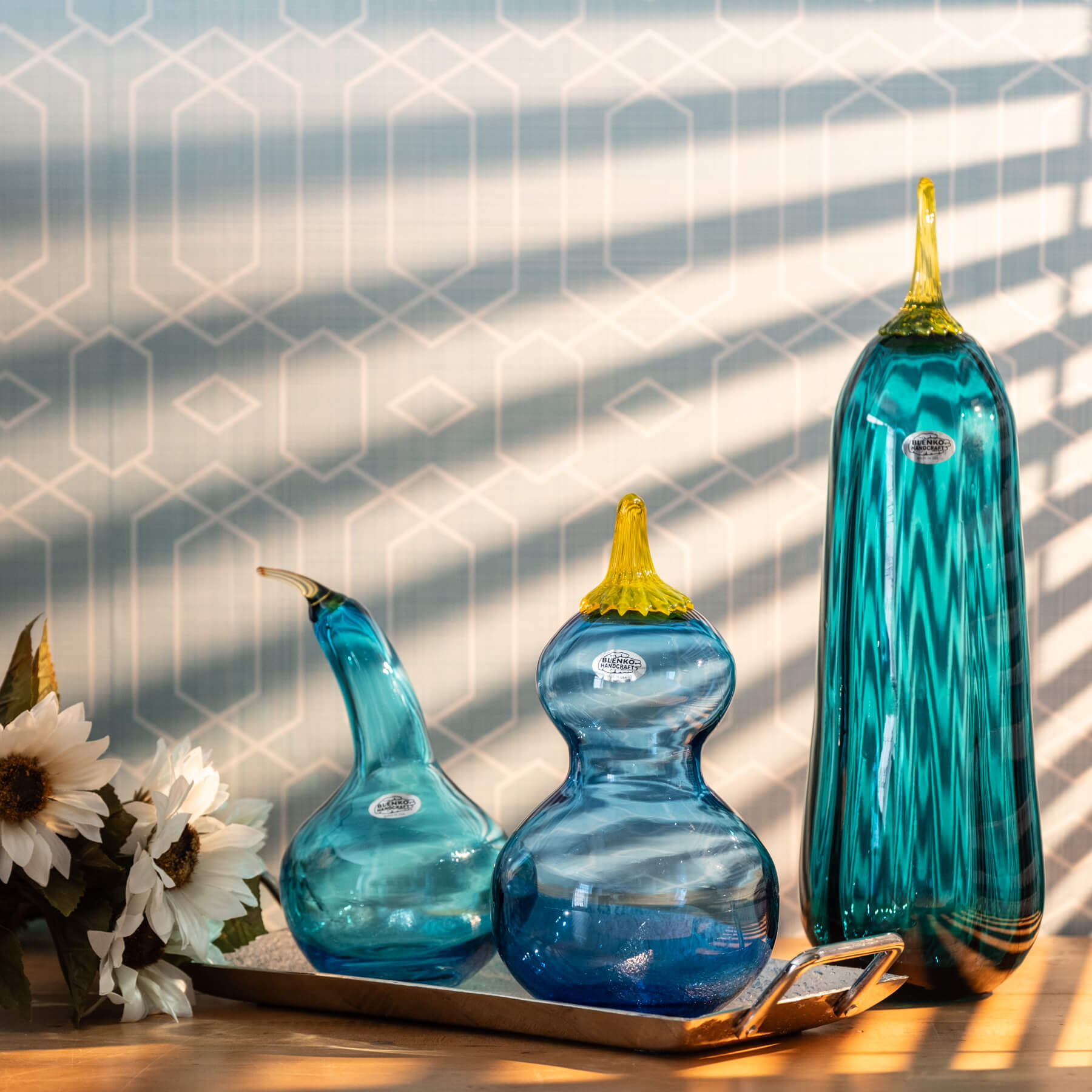 Three blue glass gourds with yellow stems on a wooden surface with a geometric patterned wall in the background.
