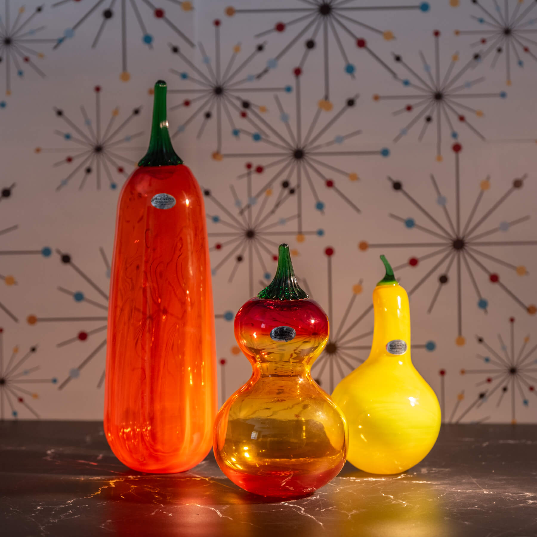 Three colorful glass gourds on a reflective surface with a patterned wall in the background.