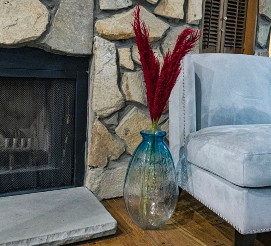Tall crystal vase with blue top containing maroon plant by a fireplace.