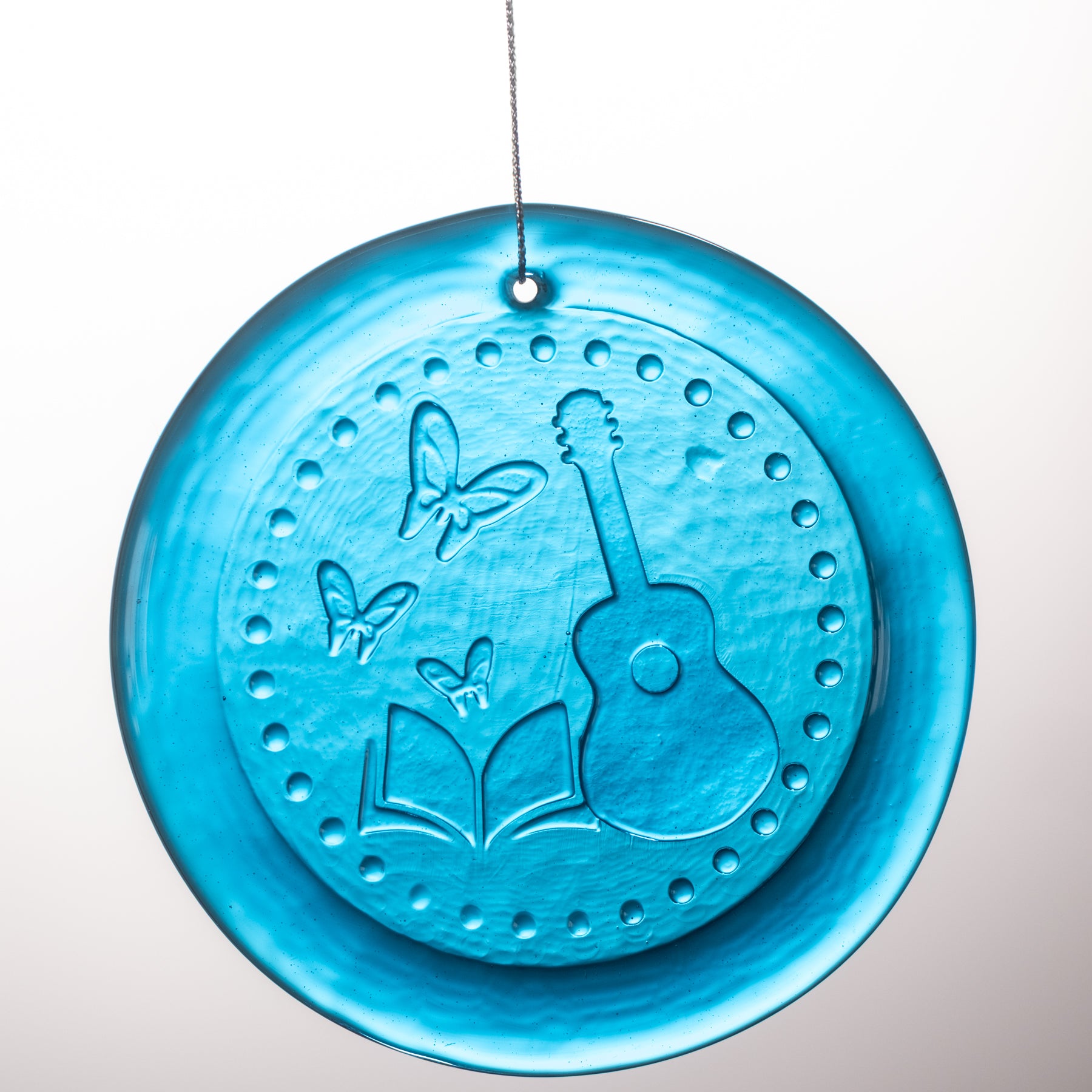 A round turquoise blue glass hanging ornament featuring embossed designs of a guitar, butterflies, an open book, and a dotted border, suspended by a thin string against a light gray gradient background.