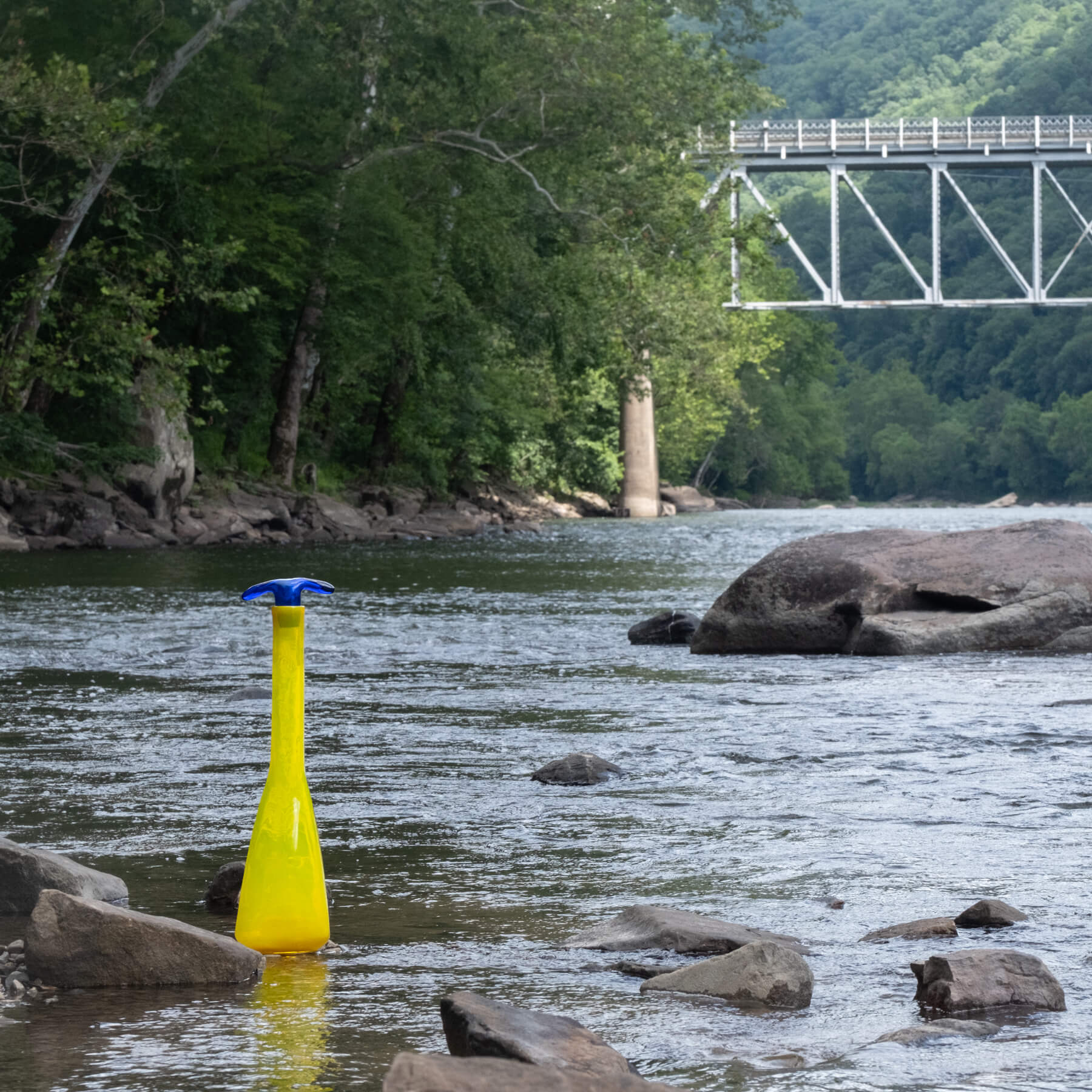 Yellow bottle with blue cap floating in a river with a bridge and trees in the background