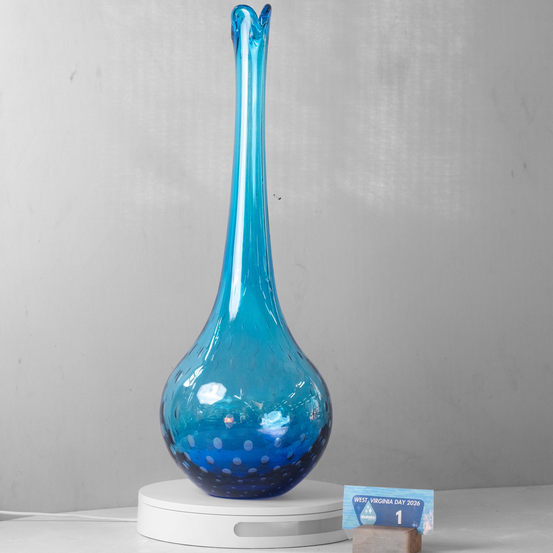 A tall, slender blue glass vase with a bulbous base and small white speckles, displayed on a white circular pedestal against a light gray wall.