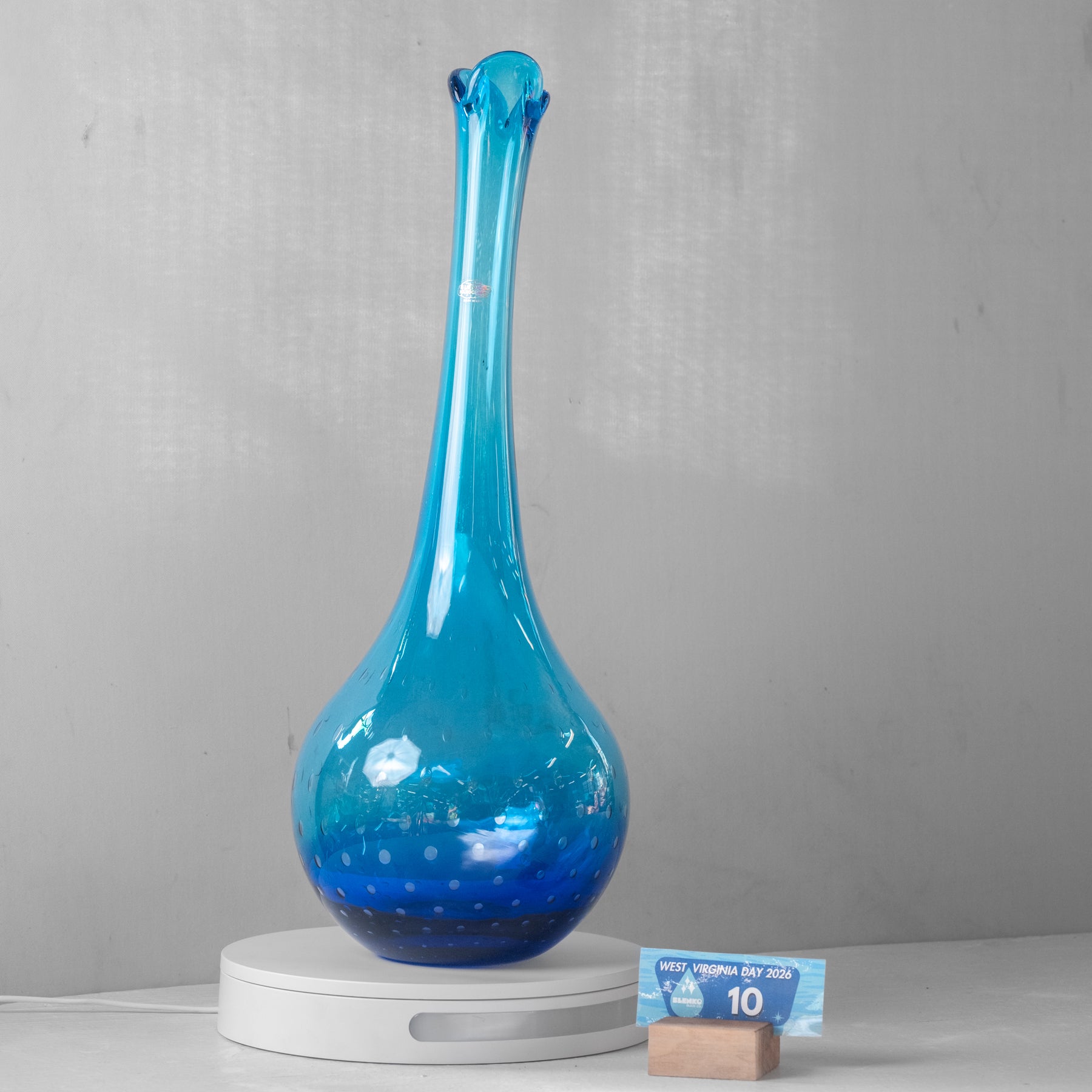 A tall, bulb-shaped blue glass decanter or vase with a narrow neck and flared rim, displayed on a white circular stand against a light gray wall. A small blue card and wooden block are placed beside the stand.