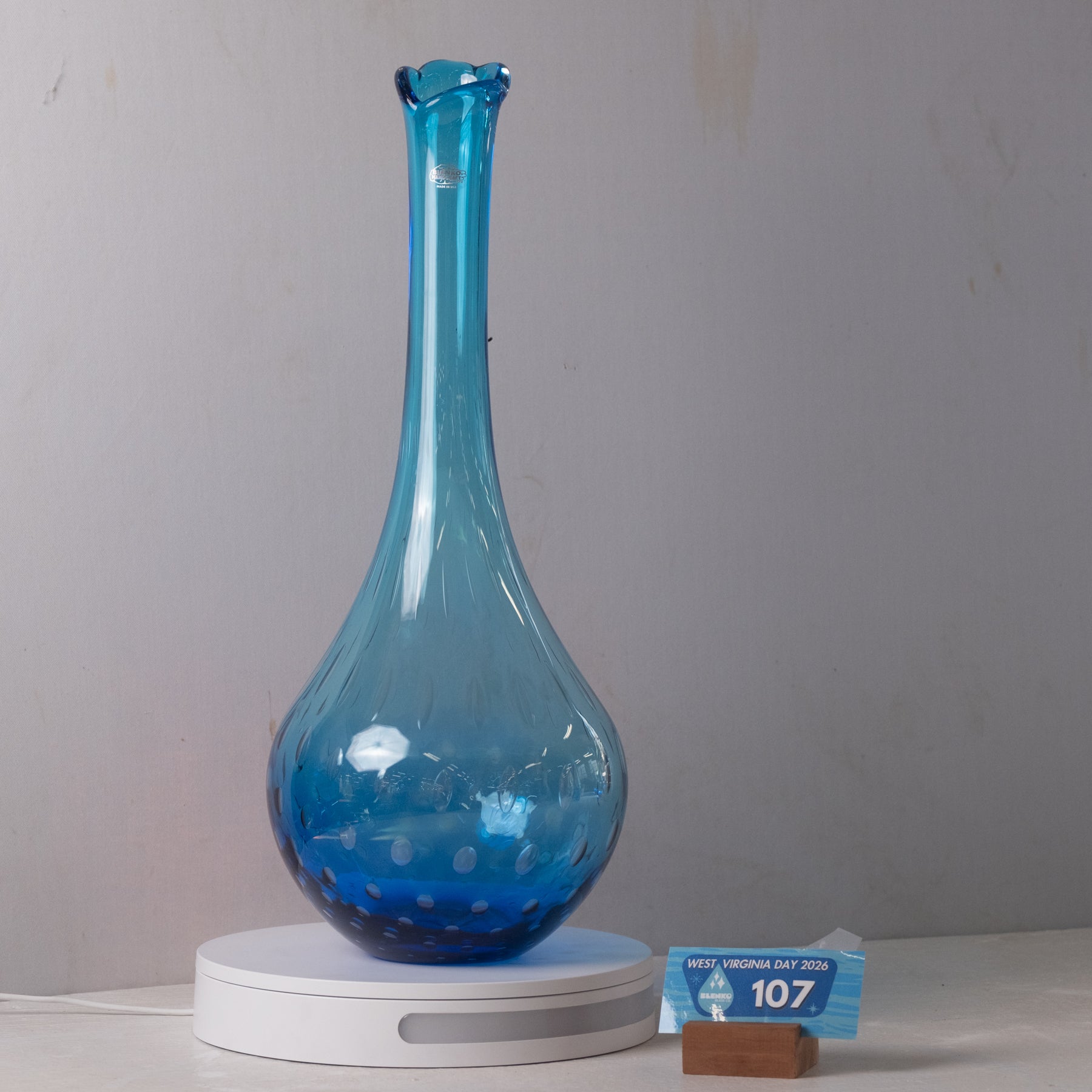 A translucent blue glass vase with a flared neck and dotted pattern near the base, displayed on a white round stand against a plain light gray wall.