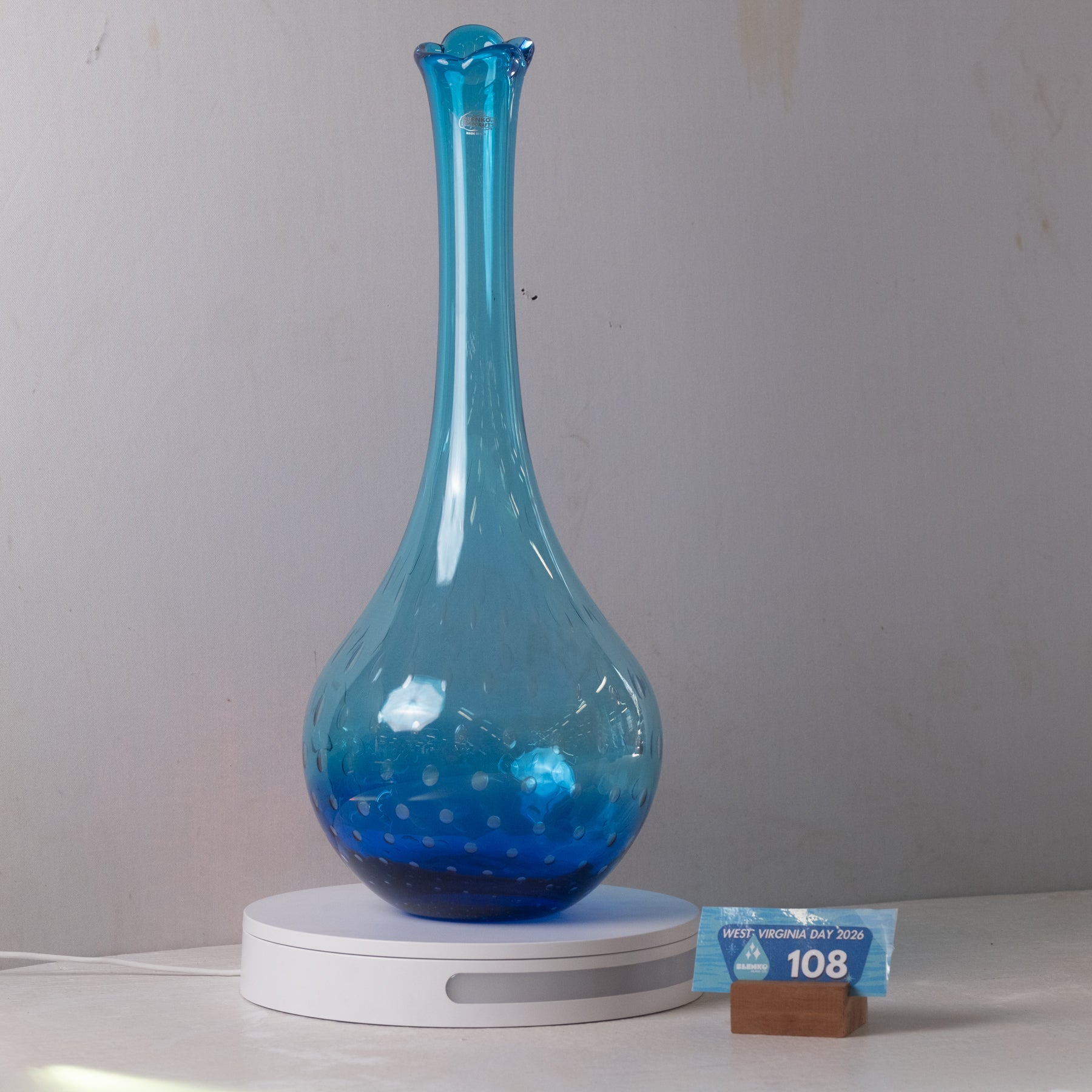 A translucent blue glass vase with a flared neck and rounded base, displayed on a white circular light box. A small blue price tag card is placed beside the base on a white surface against a plain light gray wall.