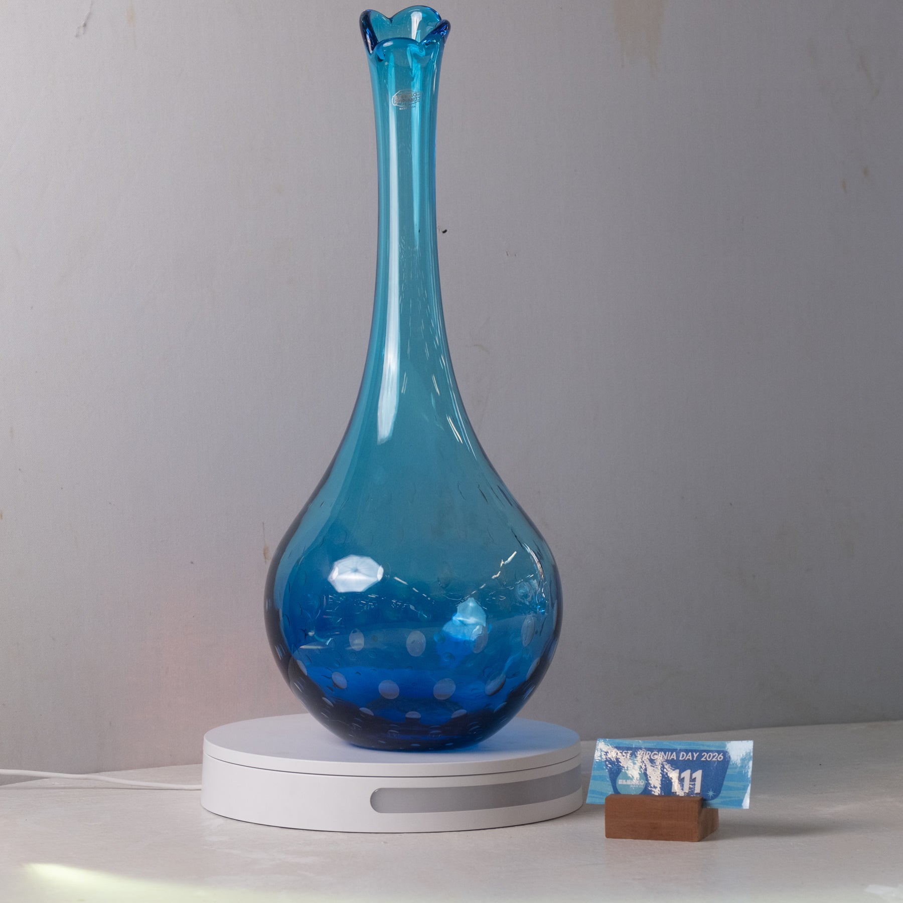 A translucent blue glass vase with a flared rim and bulbous base, displayed on a white circular stand against a plain light gray wall. A small card is propped beside the stand.