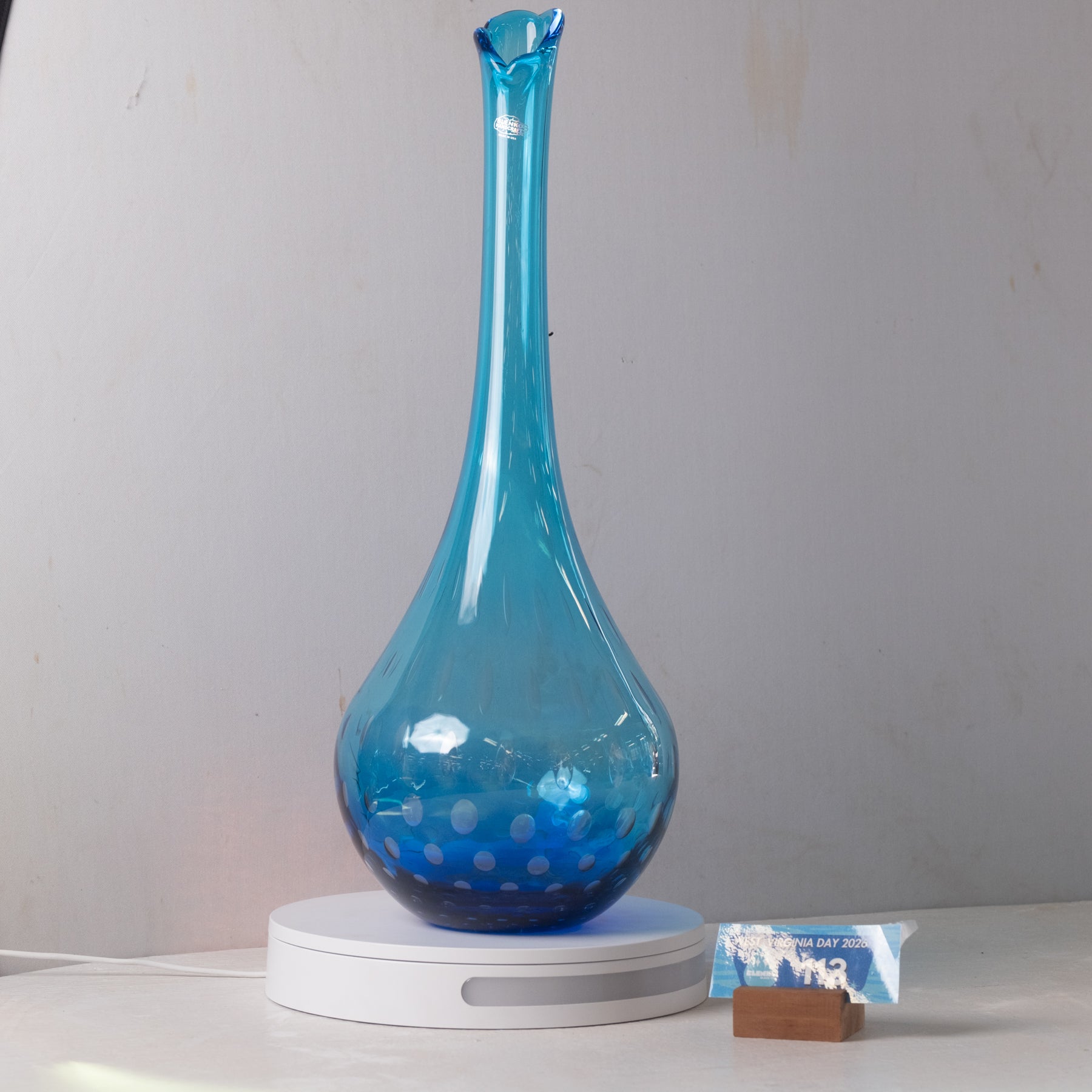A translucent blue glass vase with a flared neck and dotted pattern near the base, displayed on a white circular light box. A small wrapped item and a brown block are placed beside it on a white surface against a plain light gray wall.
