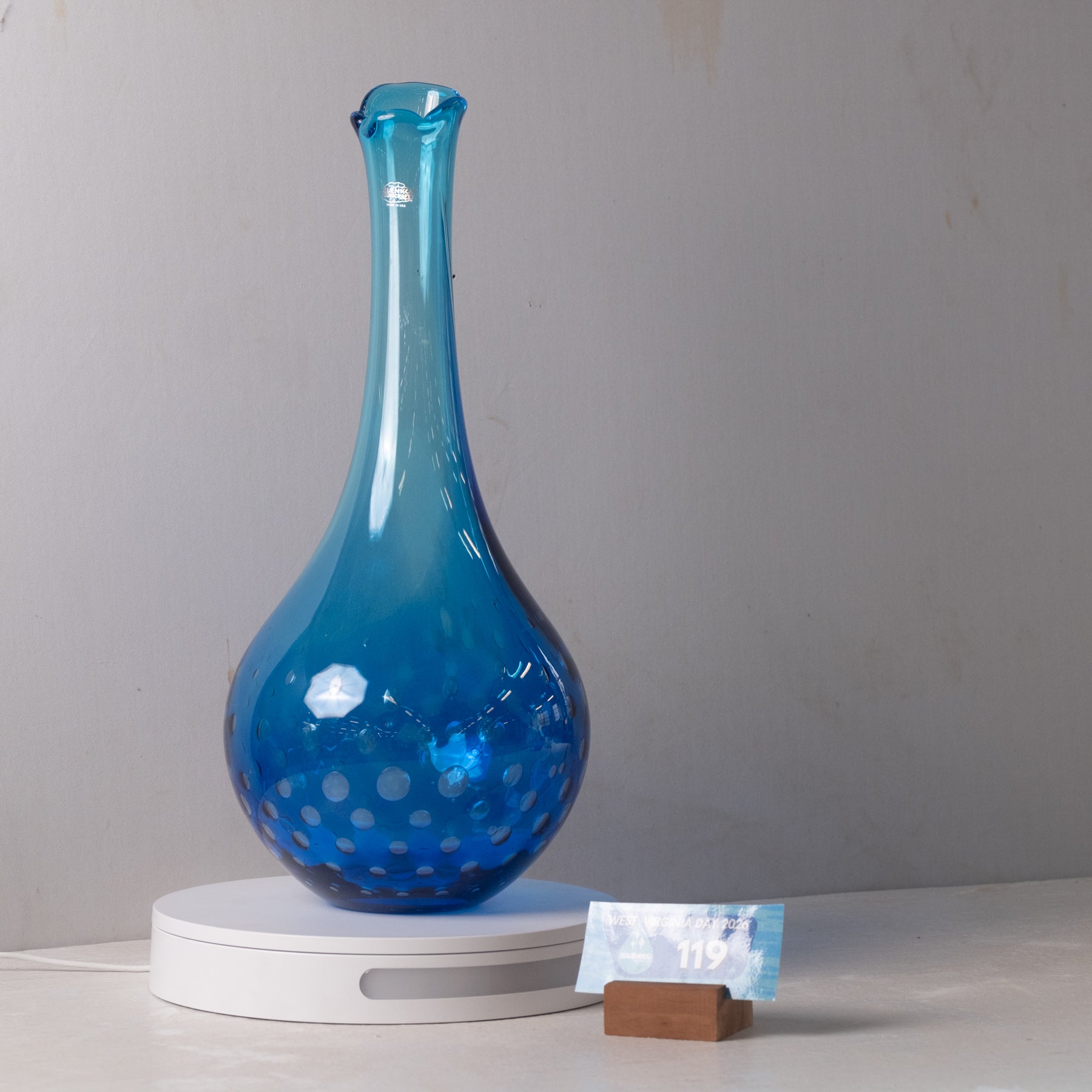 A translucent blue glass vase with a bulbous base and narrow neck, decorated with small white polka dots, displayed on a white circular stand against a light gray wall.