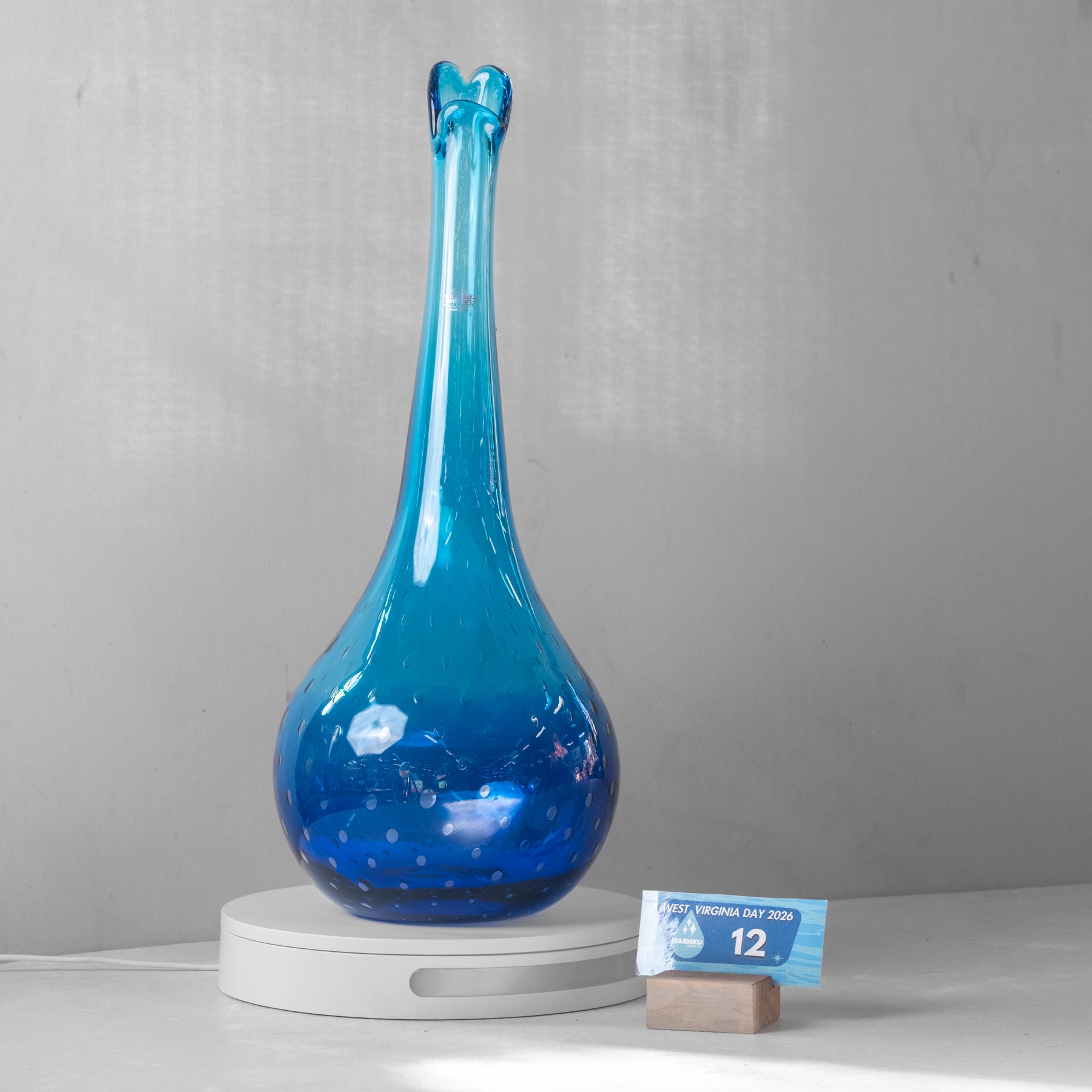 A tall, bulb-shaped blue glass vase with a narrow neck and small flared top, displayed on a white round pedestal. A small blue calendar card stands beside it on a light surface against a gray wall.