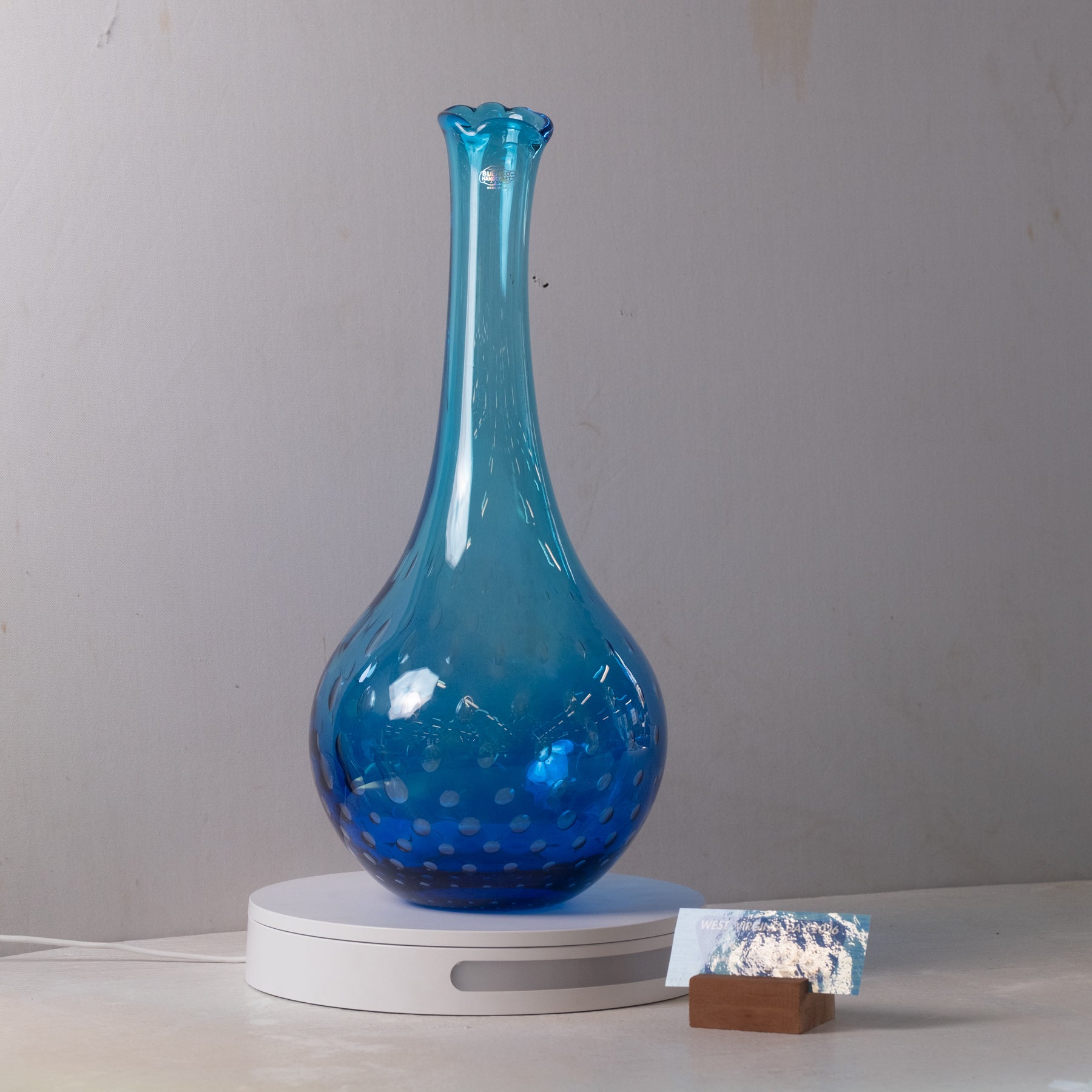 A translucent blue glass vase with a flared neck and dotted pattern near the base, displayed on a white circular stand against a plain light gray wall.
