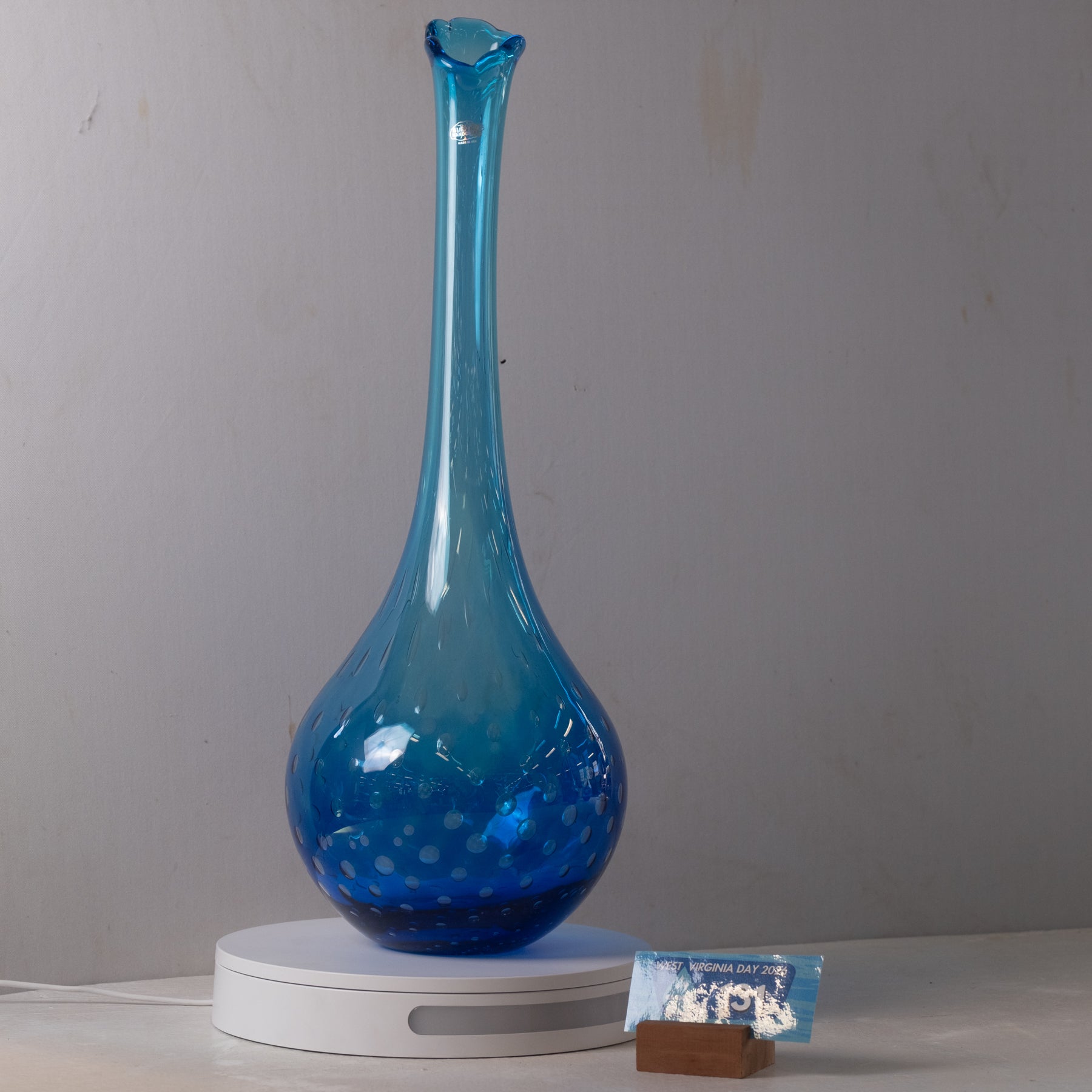 A translucent blue glass vase with a long, narrow neck and a rounded base speckled with small bubbles, displayed on a white circular stand against a plain light gray wall.