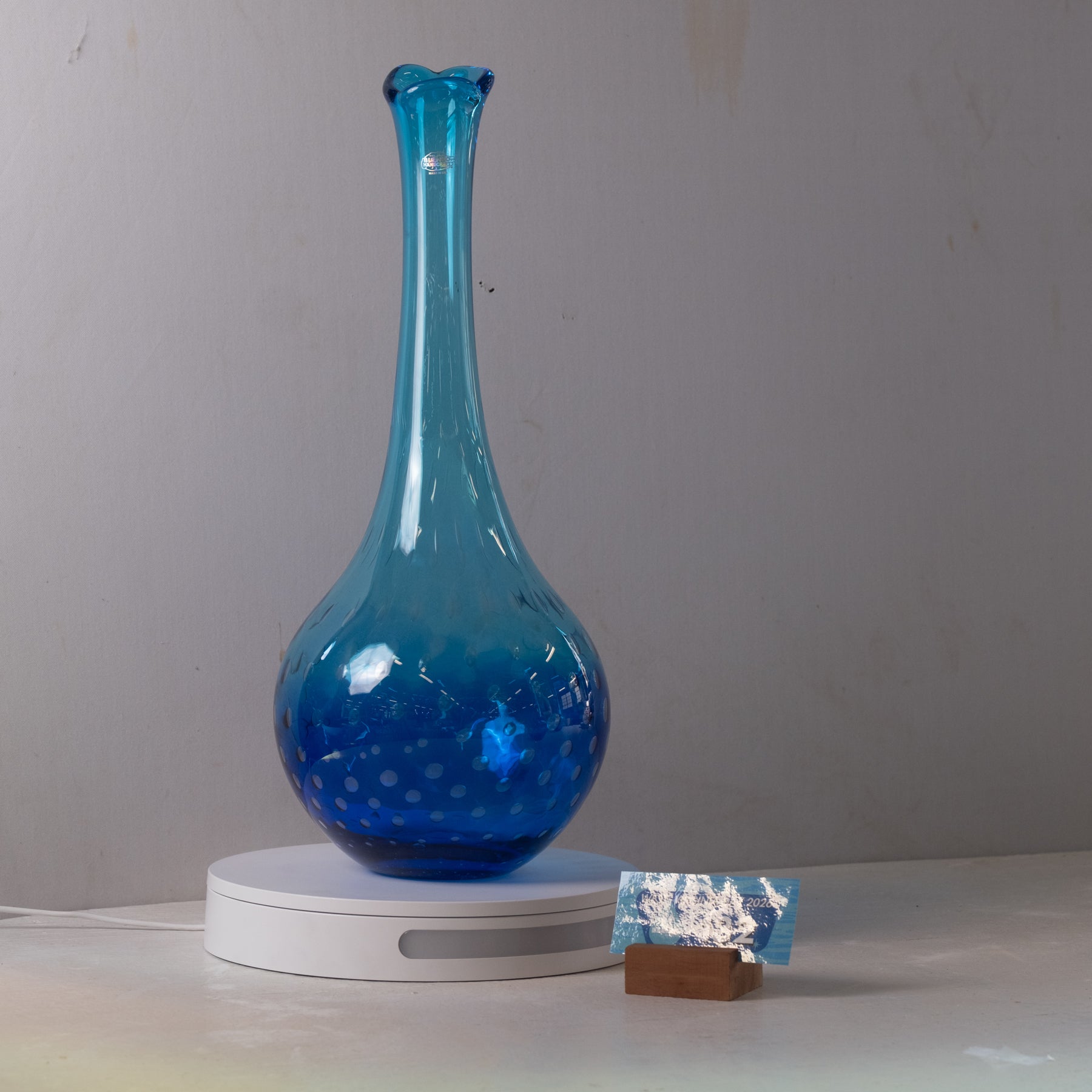 A translucent blue glass vase with a flared neck and dotted pattern, displayed on a white circular base against a plain gray wall.