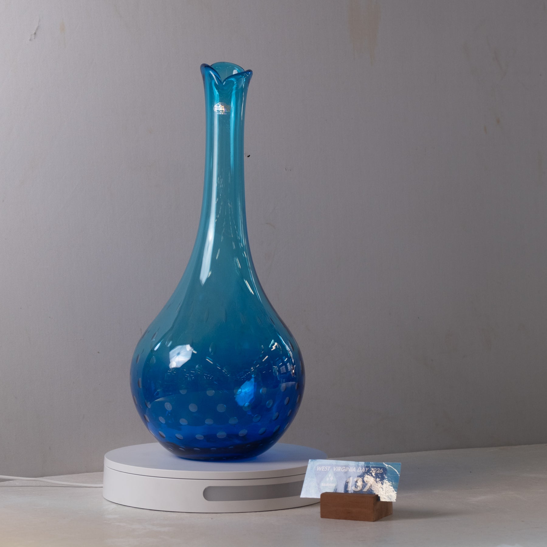 A translucent blue glass vase with a flared neck and dotted pattern, displayed on a white circular light box against a plain gray wall.