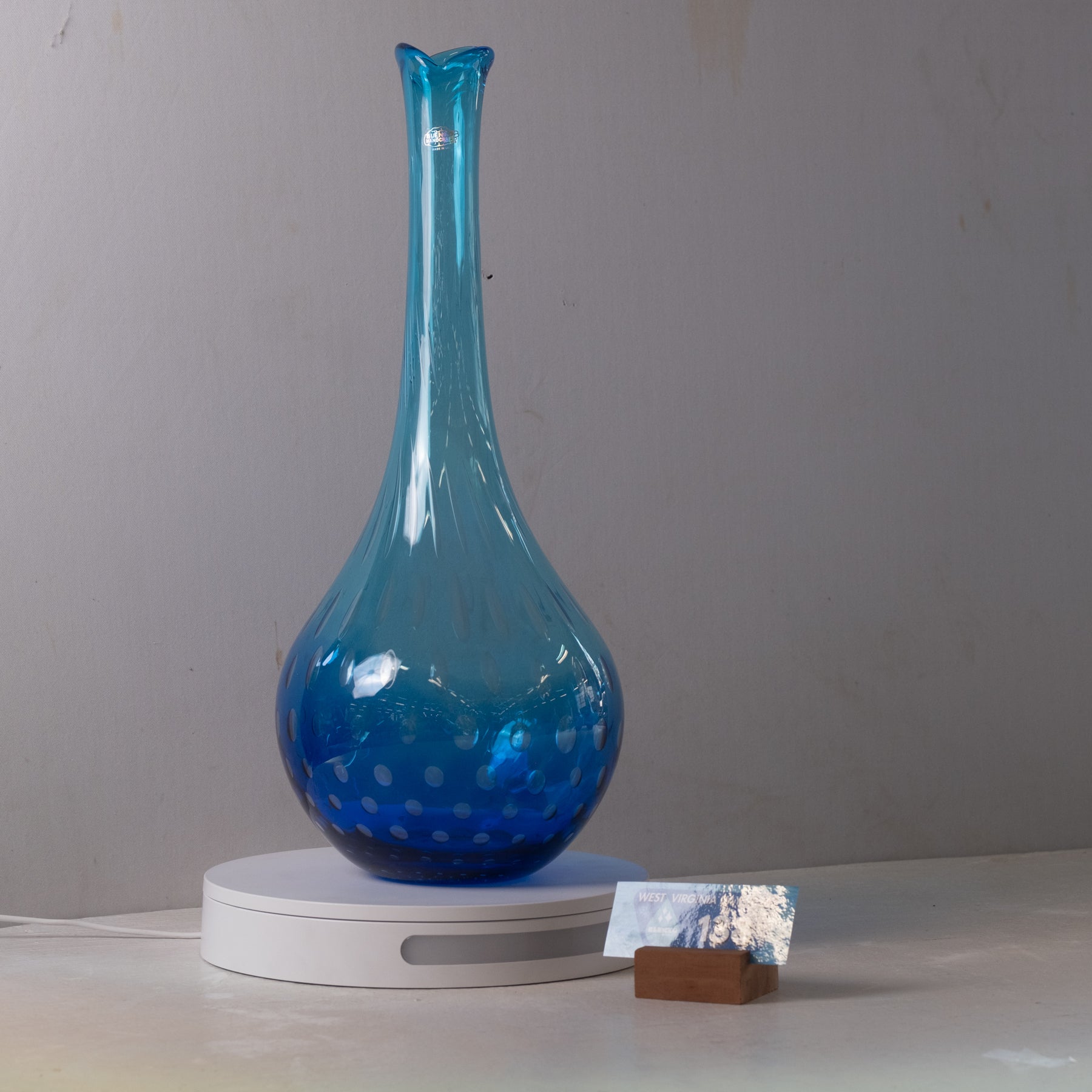 A translucent blue glass vase with a flared neck and dotted pattern near the base, displayed on a white circular stand against a plain gray wall.