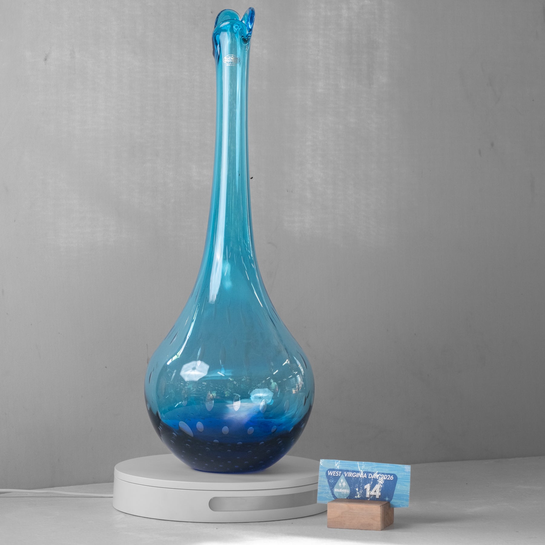 A tall, narrow blue glass vase with a bulbous base and a flared rim, displayed on a white round pedestal against a gray wall. A small blue card on a wooden block sits beside the pedestal.