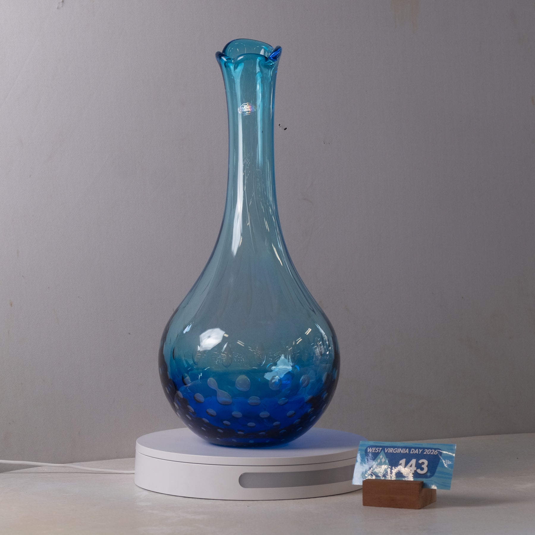 A translucent blue glass vase with a flared rim and speckled pattern near the base, displayed on a white circular light box against a plain gray wall.