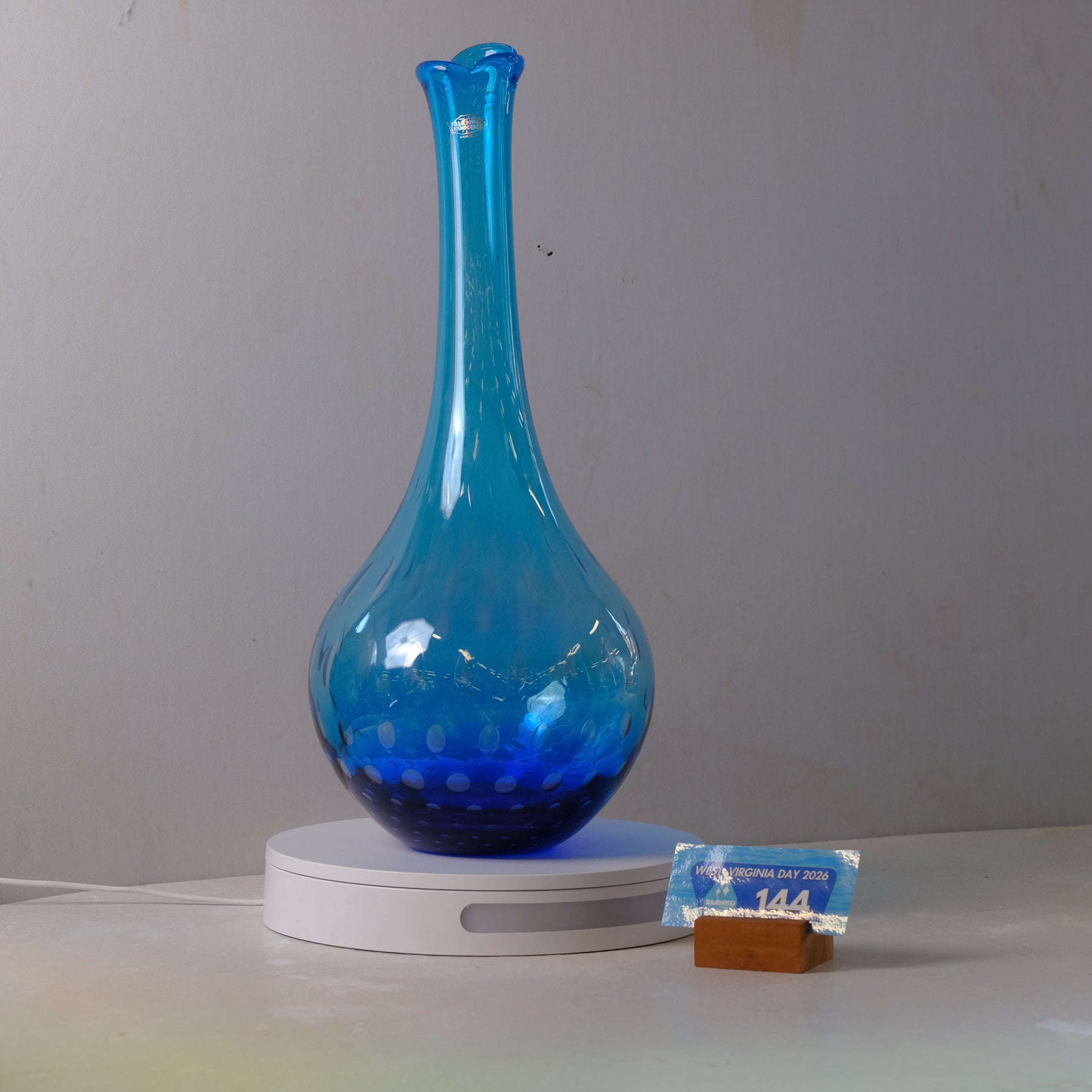 A translucent blue glass vase with a flared neck and rounded base, displayed on a white circular light box. A small price tag card is visible next to the base on a light-colored surface against a plain gray wall.