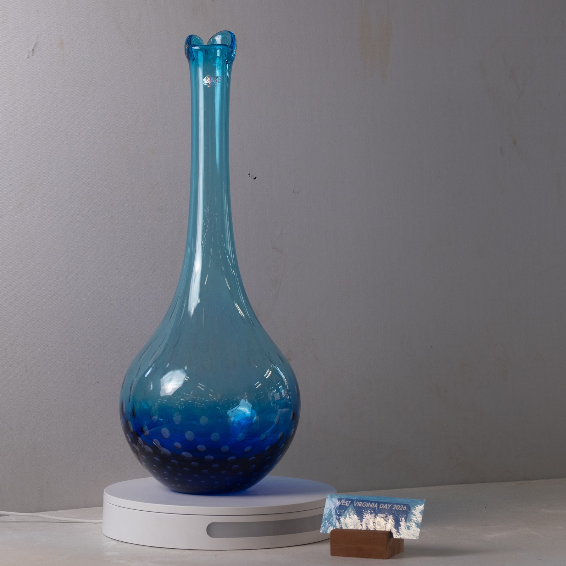 A translucent blue glass vase with a flared neck and dotted texture, displayed on a white circular light box against a plain gray wall.
