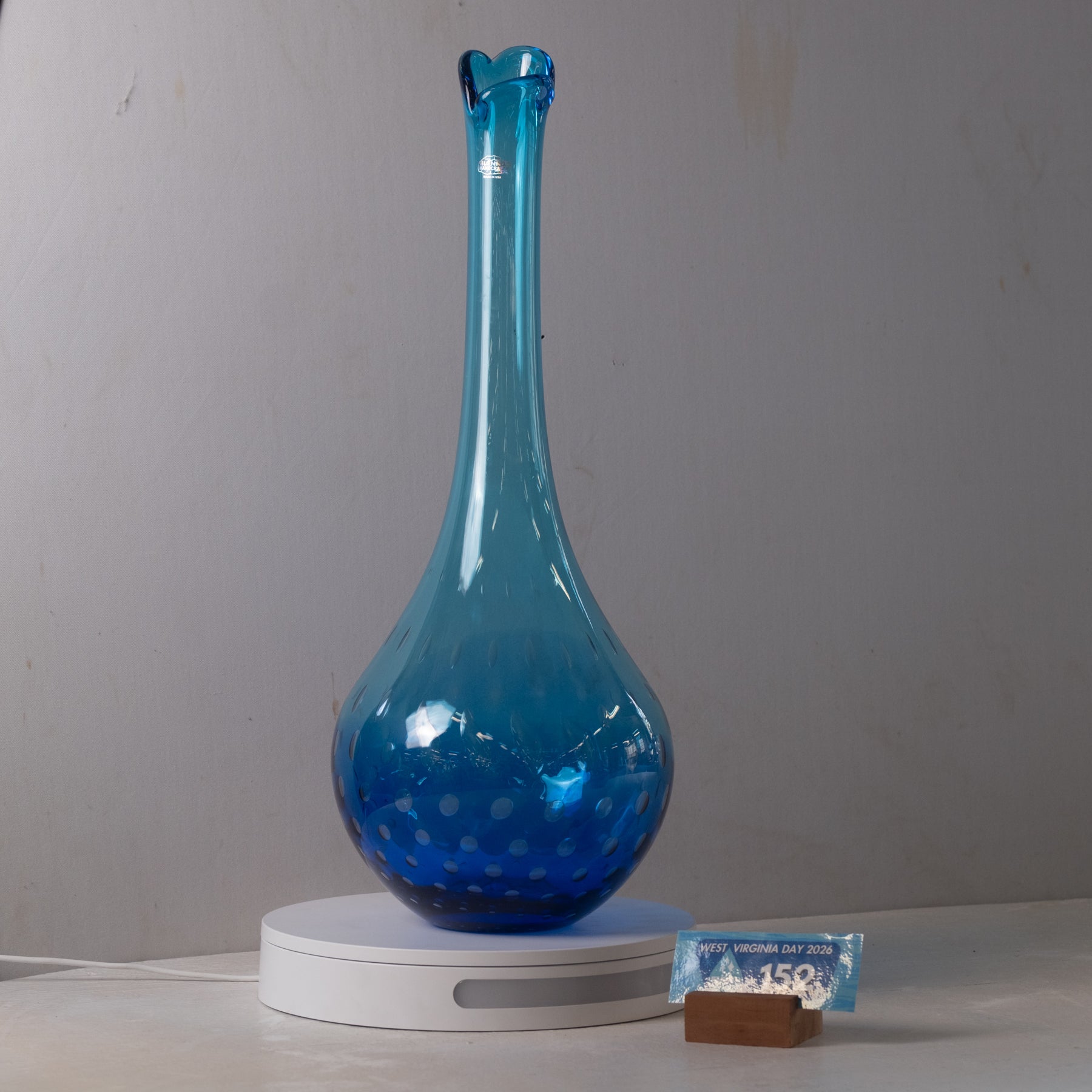 A translucent blue glass vase with a flared rim and dotted pattern near the base, displayed on a white circular stand against a plain light gray wall.