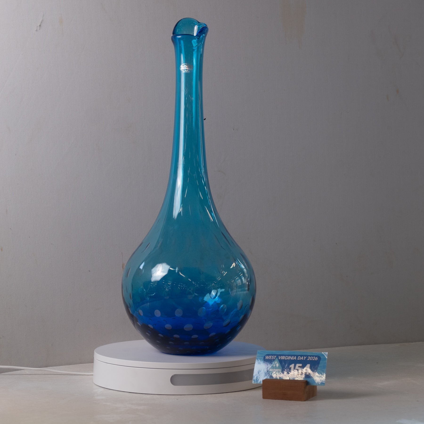 A translucent blue glass decanter with a bulbous body and narrow neck, displayed on a white circular base against a plain gray wall.
