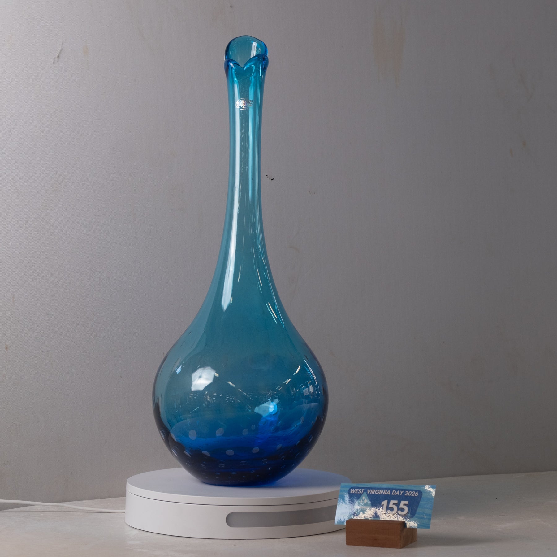 A translucent blue glass vase with a long, flared neck and bulbous base, displayed on a white circular light box. A small price tag is visible in front on a light gray background.