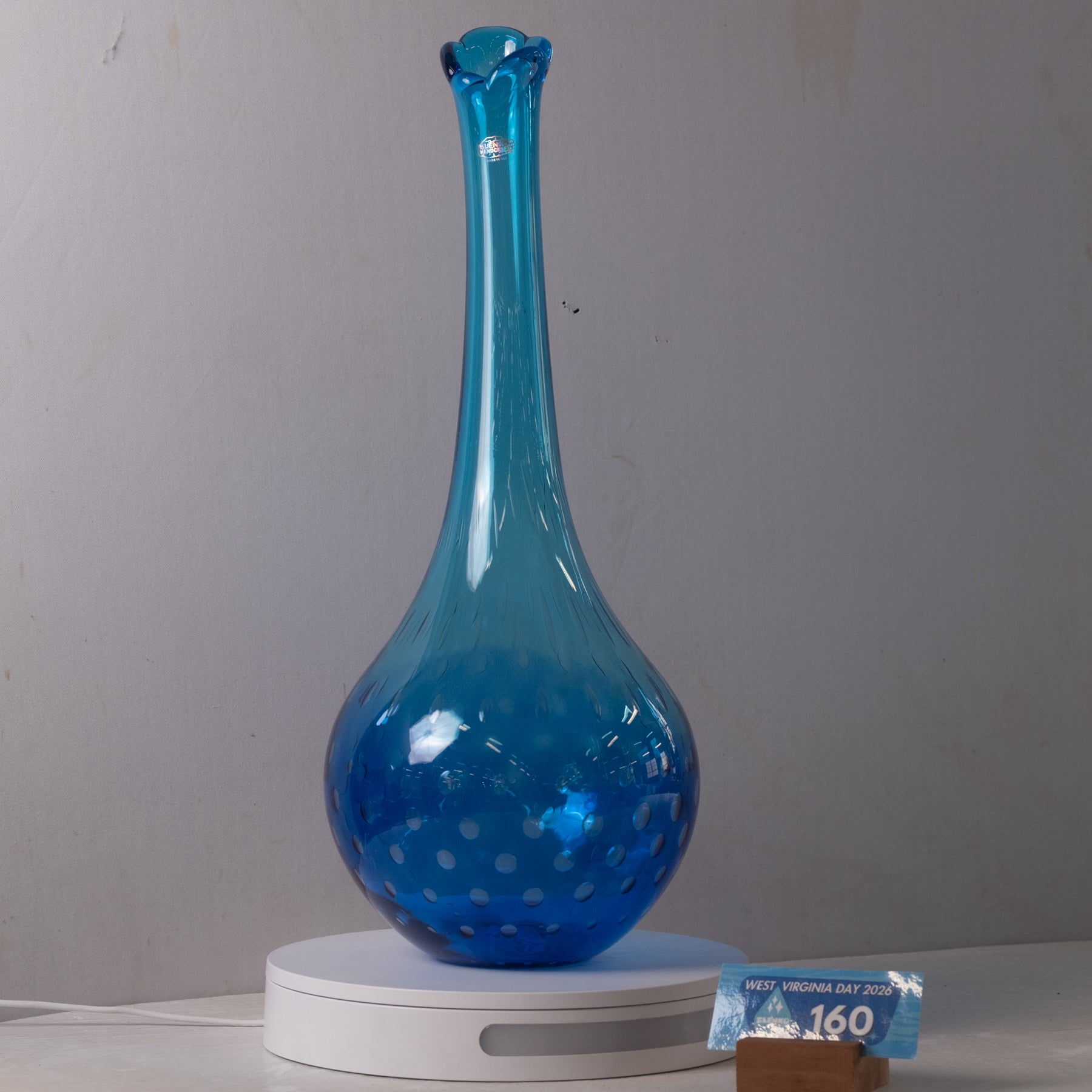 A blue glass decanter with a long neck and dotted pattern, displayed on a white circular scale against a gray wall.