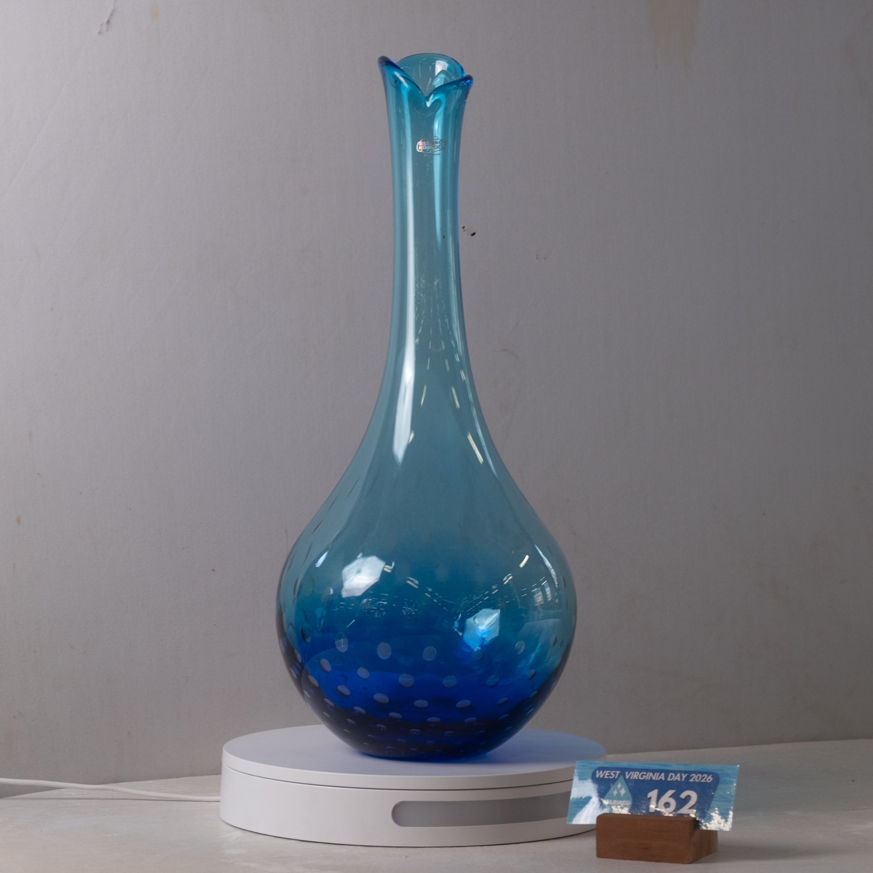 A blue glass decanter with a flared spout and rounded body, displayed on a white circular light box against a plain light gray wall.