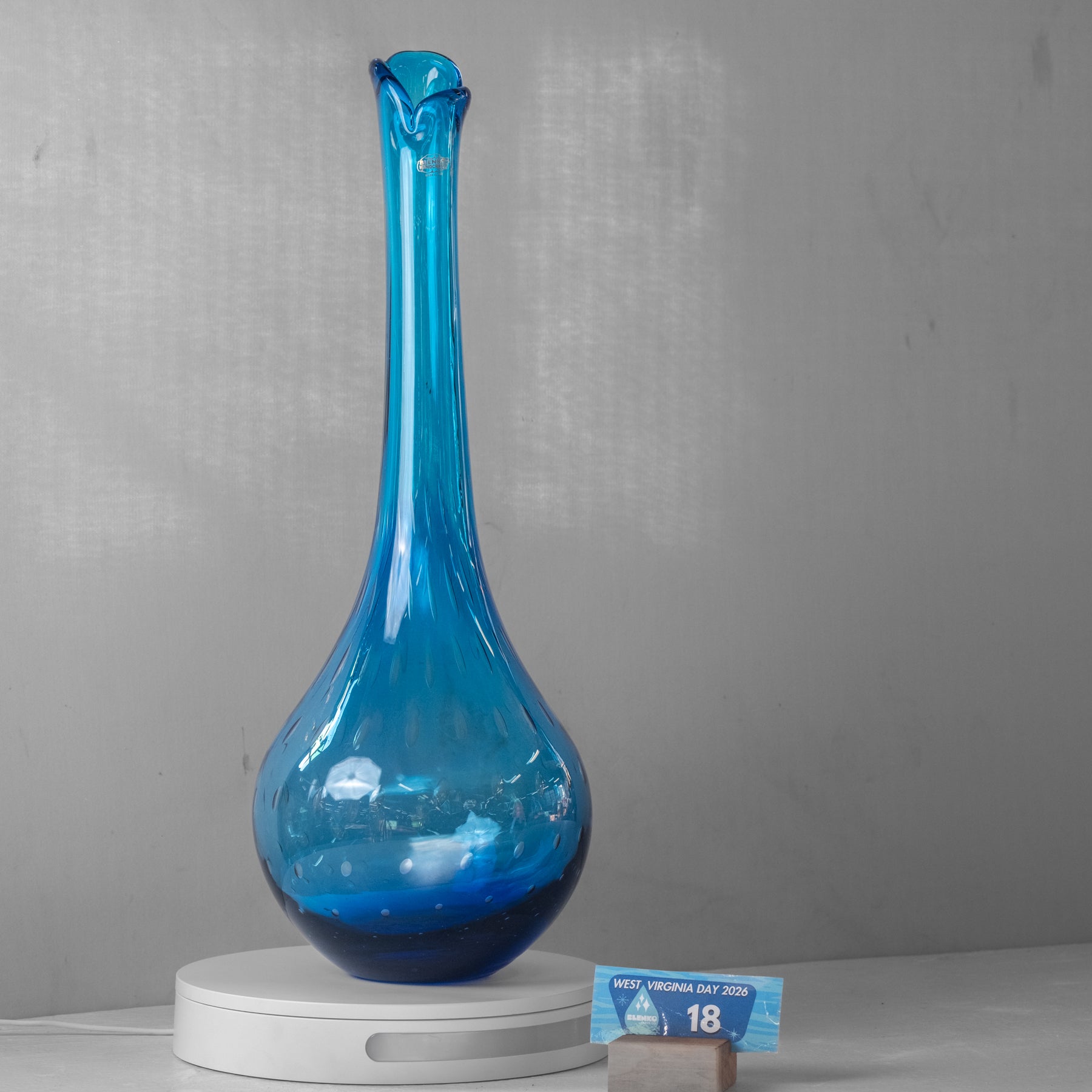 A translucent cobalt blue glass vase with a long, flared neck and bulbous base, displayed on a white circular pedestal against a gray wall. A small blue sign is visible at the bottom right.