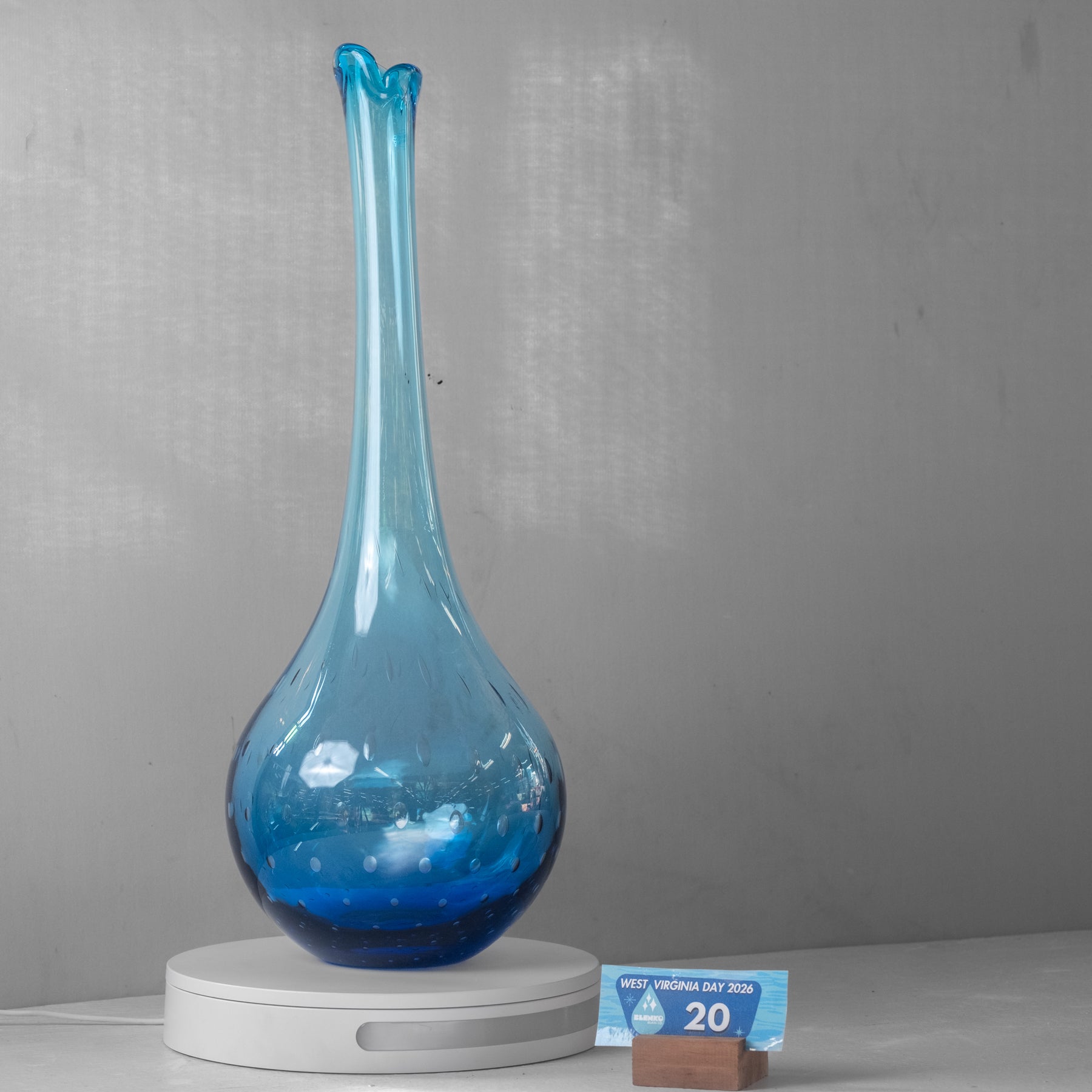 A blue glass vase with a long, narrow neck and a bulbous base, displayed on a white circular stand against a gray wall. A small sign in front reads 'WATER VALENCIA DAY 2026' and '20'.