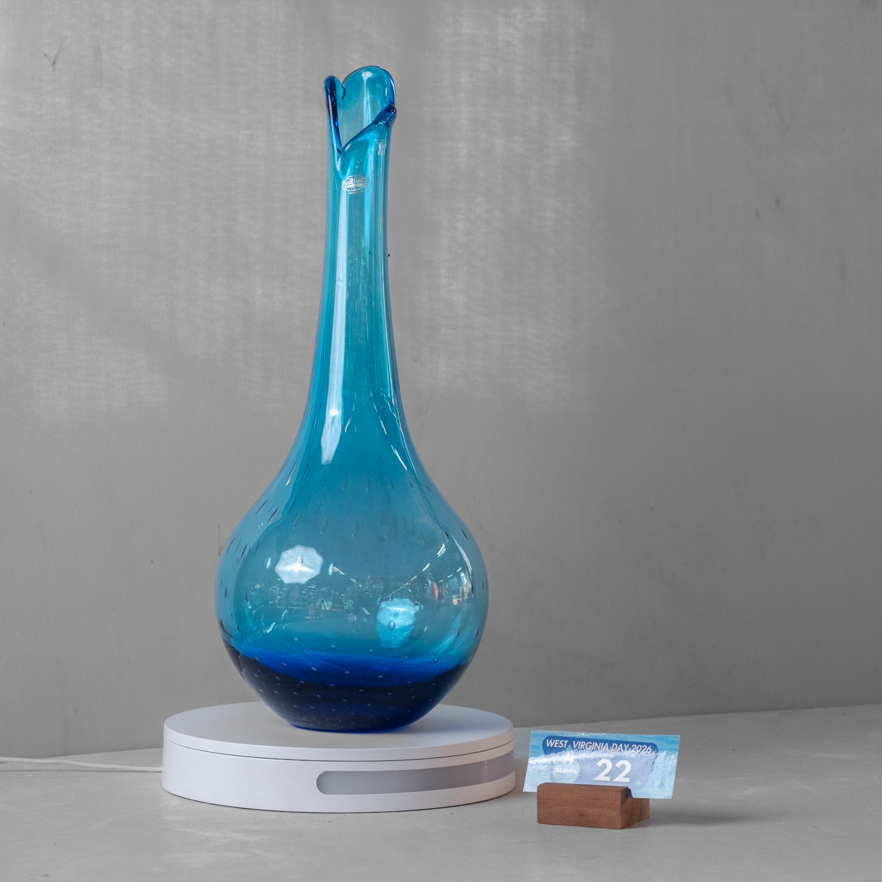 A translucent blue glass vase with a flared rim and narrow neck, displayed on a white circular base against a light gray wall. A small blue price tag card and wooden stand sit beside the base.