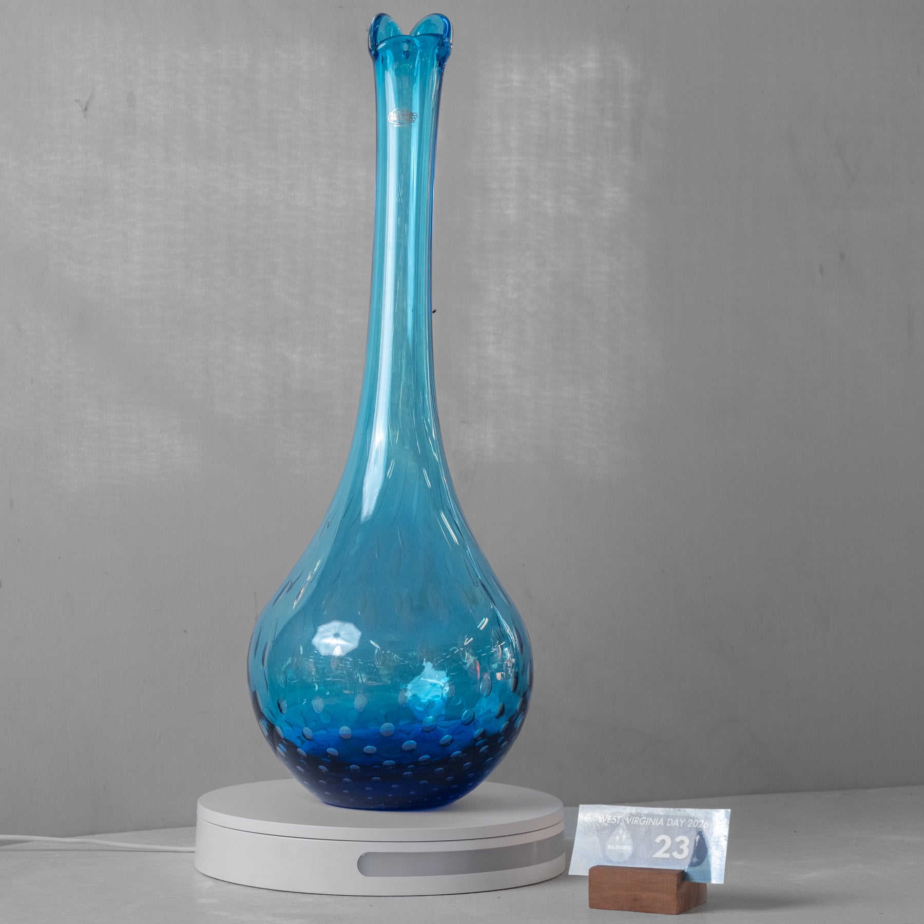 A blue glass vase with a flared neck and textured bubble pattern, displayed on a white circular stand against a gray background.