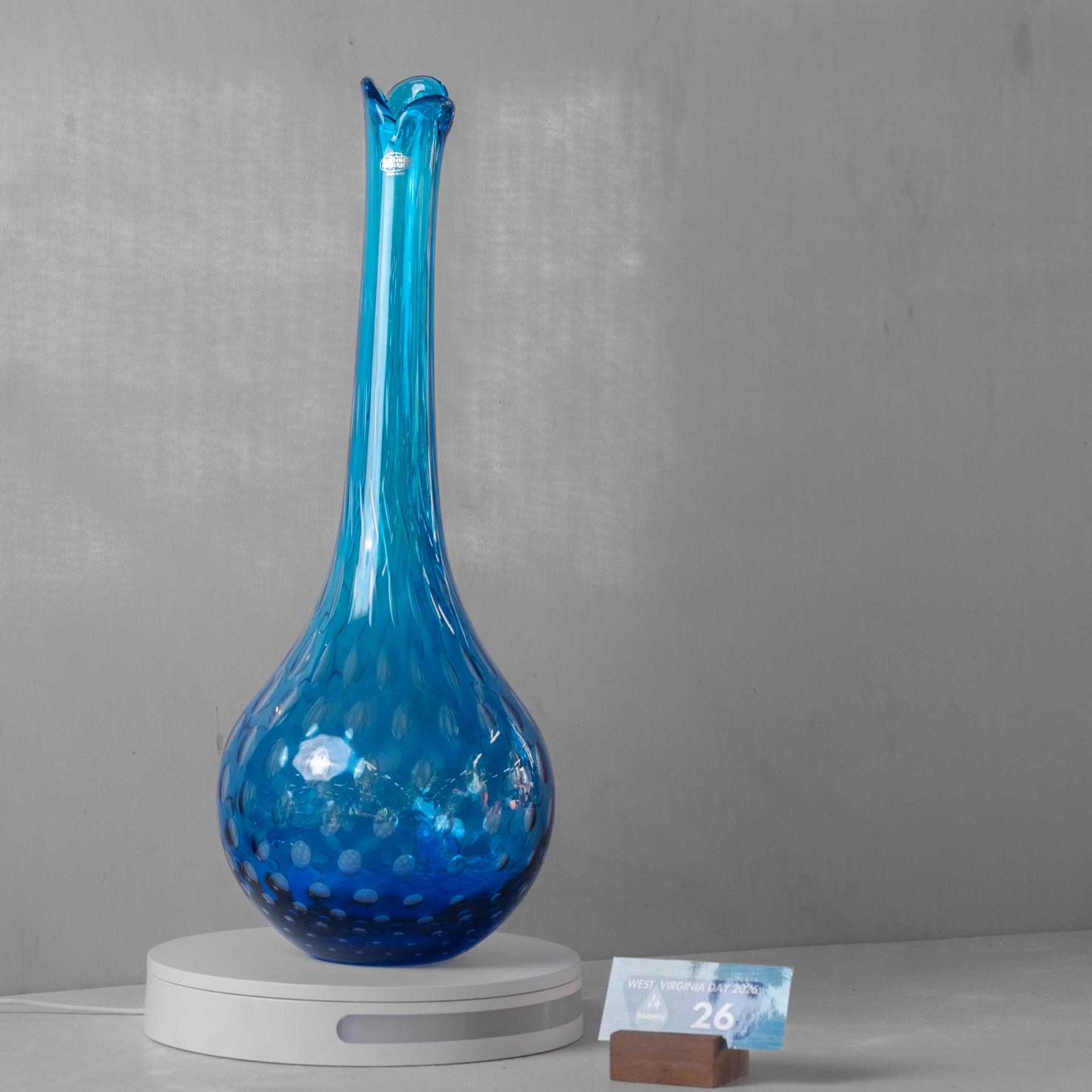 A tall, bulb-shaped blue glass vase with a narrow neck and a hammered texture, displayed on a white circular scale with a small price tag in front, against a gray wall.
