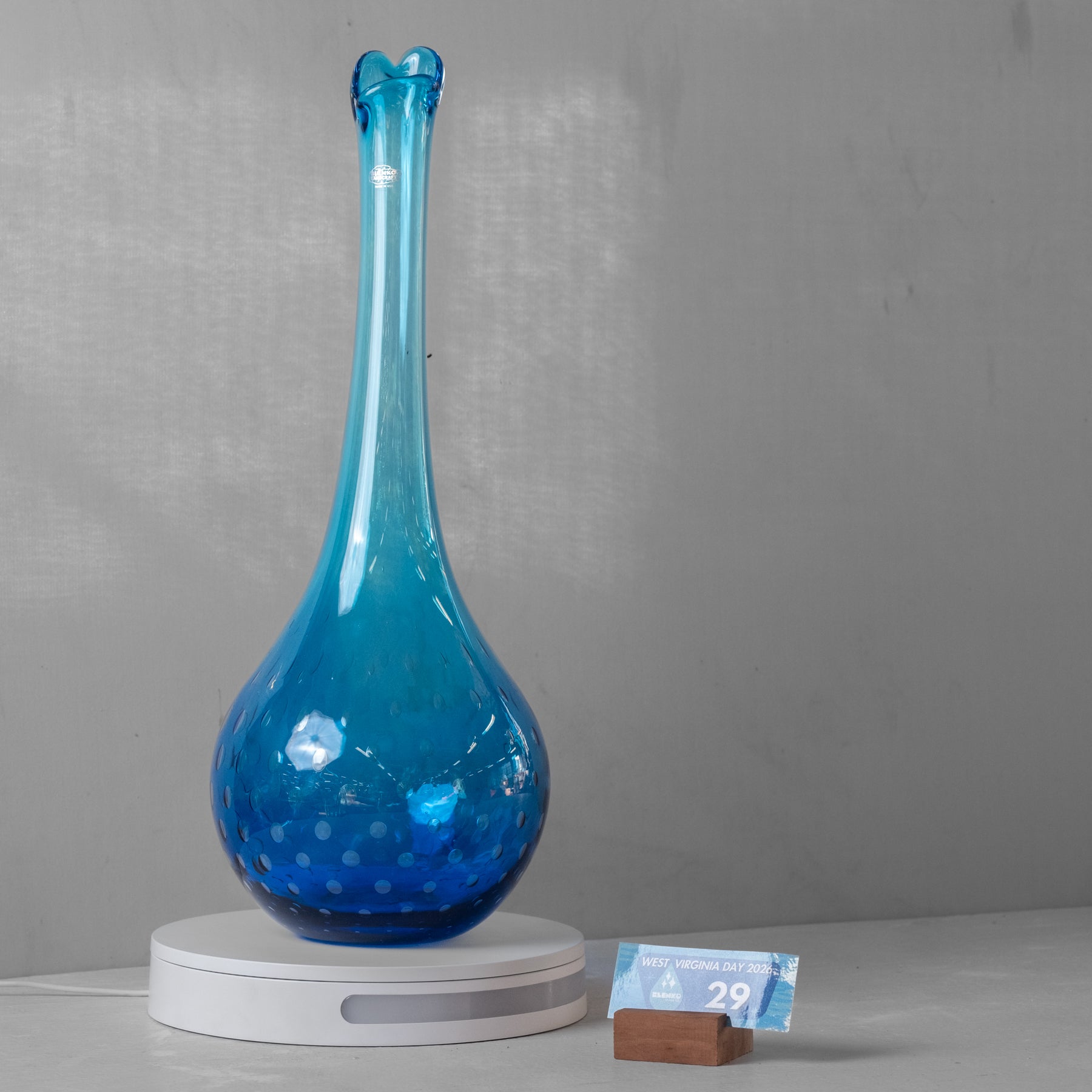 A tall, slender blue glass vase with a flared rim and dotted texture on the body, displayed on a white circular base against a gray wall.