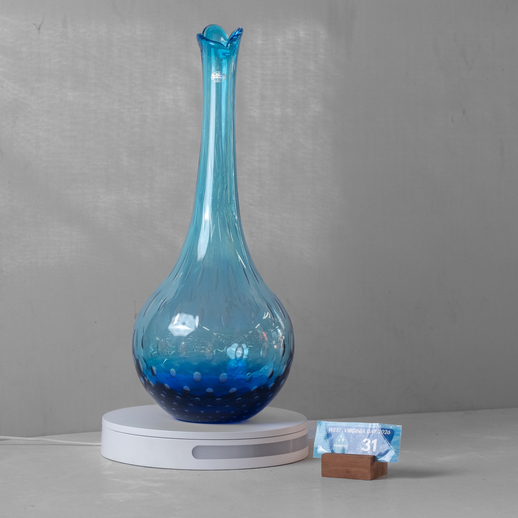A blue glass vase with a flared neck and bubble pattern, displayed on a white circular charging base with a small digital clock next to it.