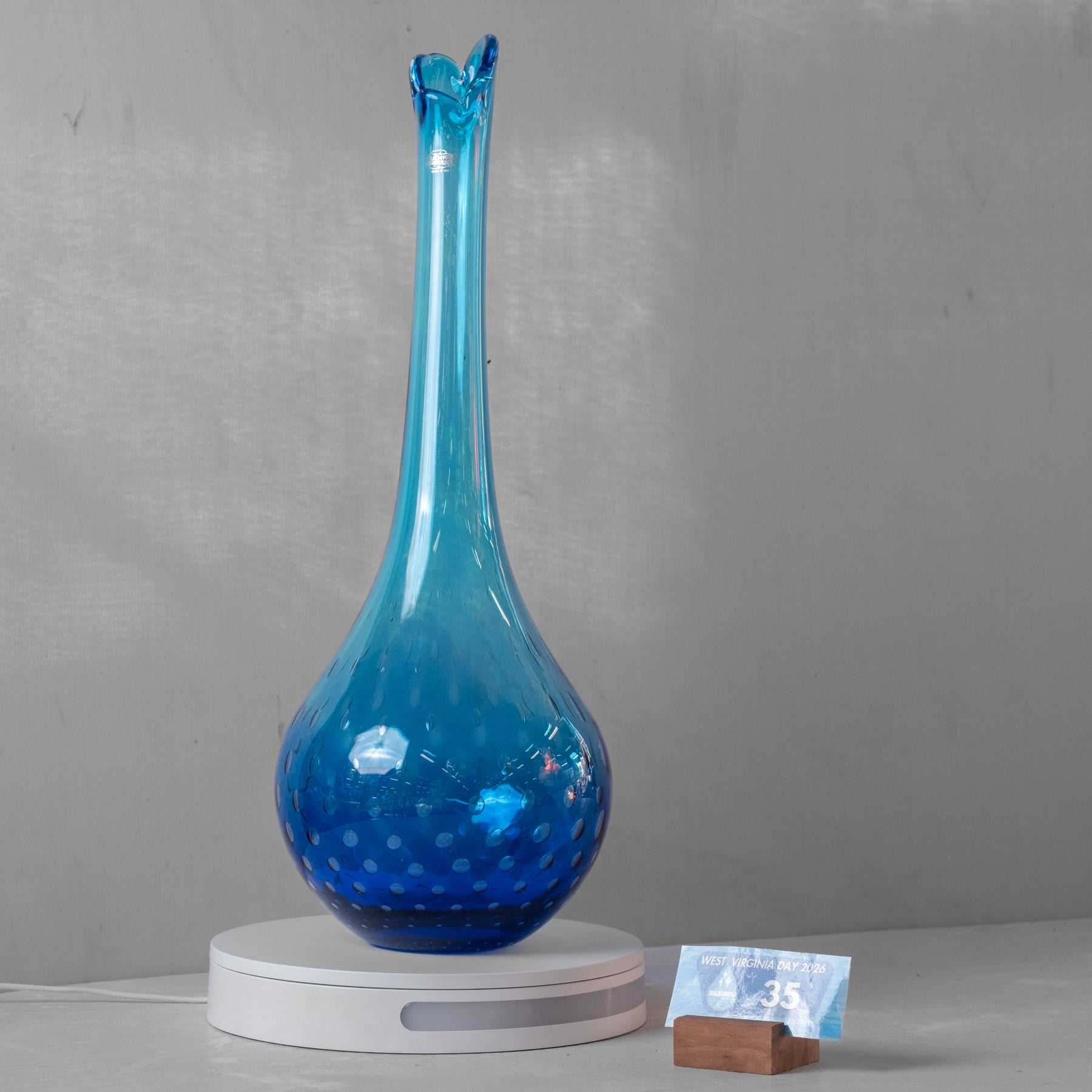 A tall, slender blue glass vase with a flared rim and bubble-patterned base, displayed on a white circular stand against a gray wall.