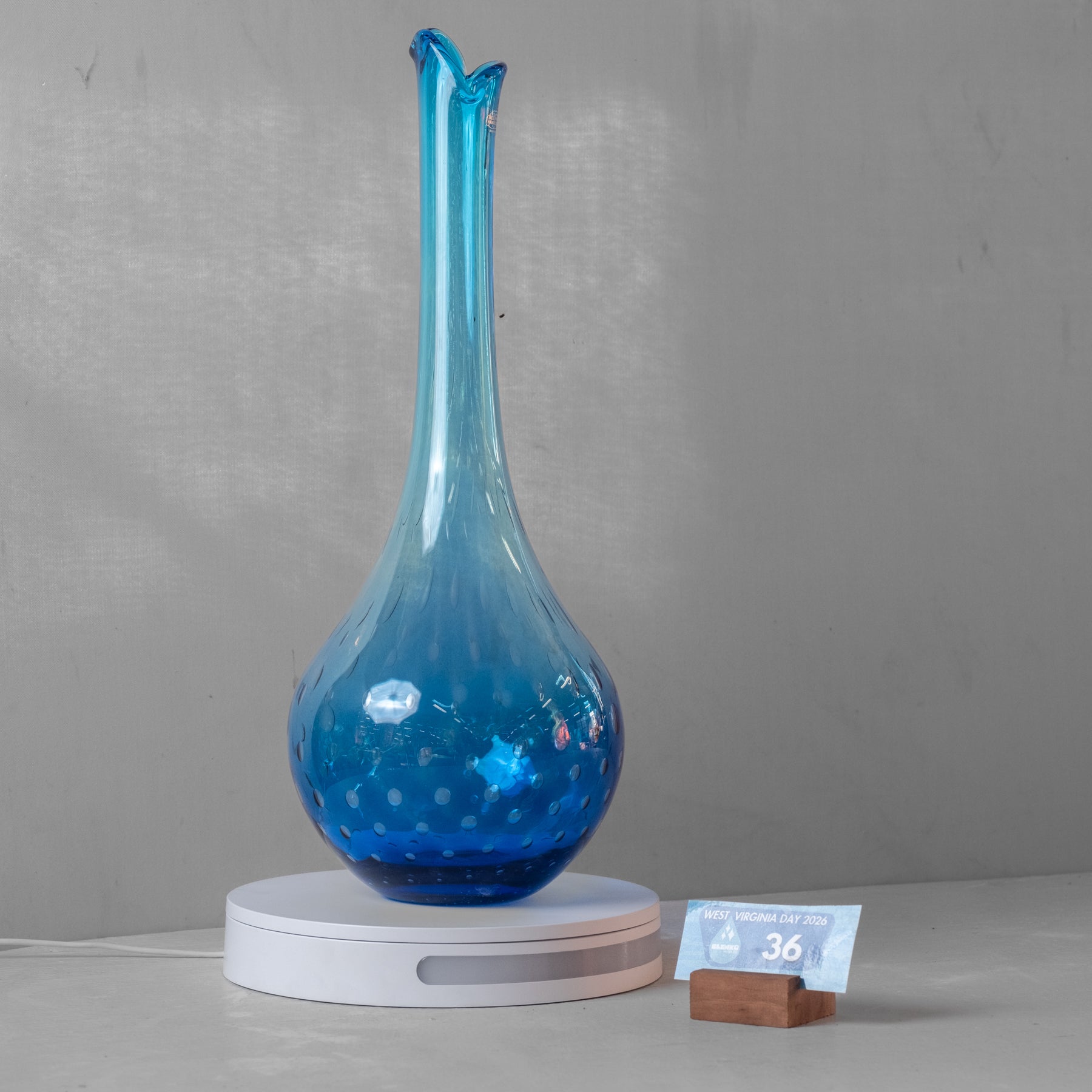 A tall, narrow blue glass vase with a bulbous base and small hand-painted fish details, displayed on a white circular light box with a small price tag in front, set against a gray background.