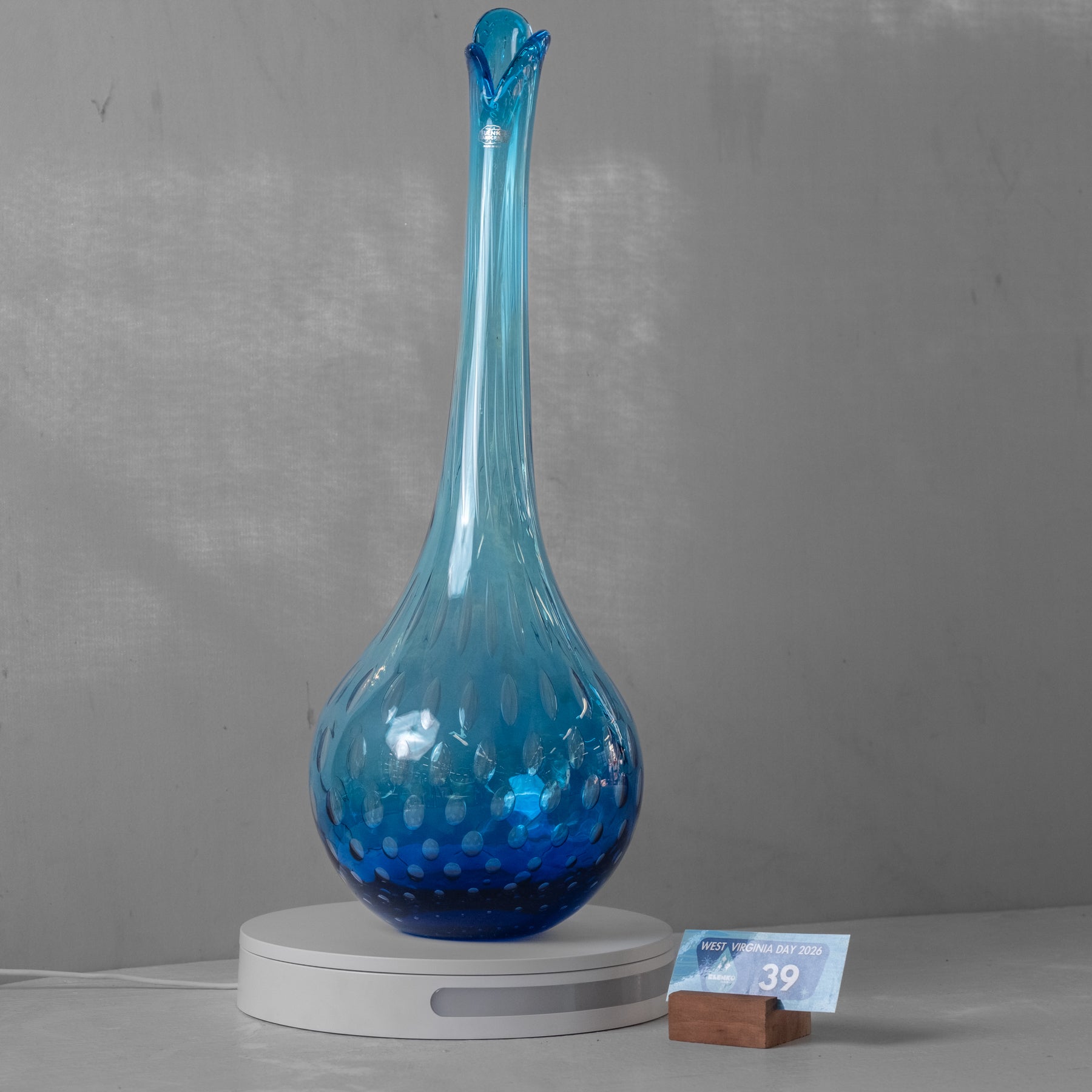 A tall, slender blue glass vase with a flared neck and textured bubble pattern, displayed on a white circular stand against a gray wall.