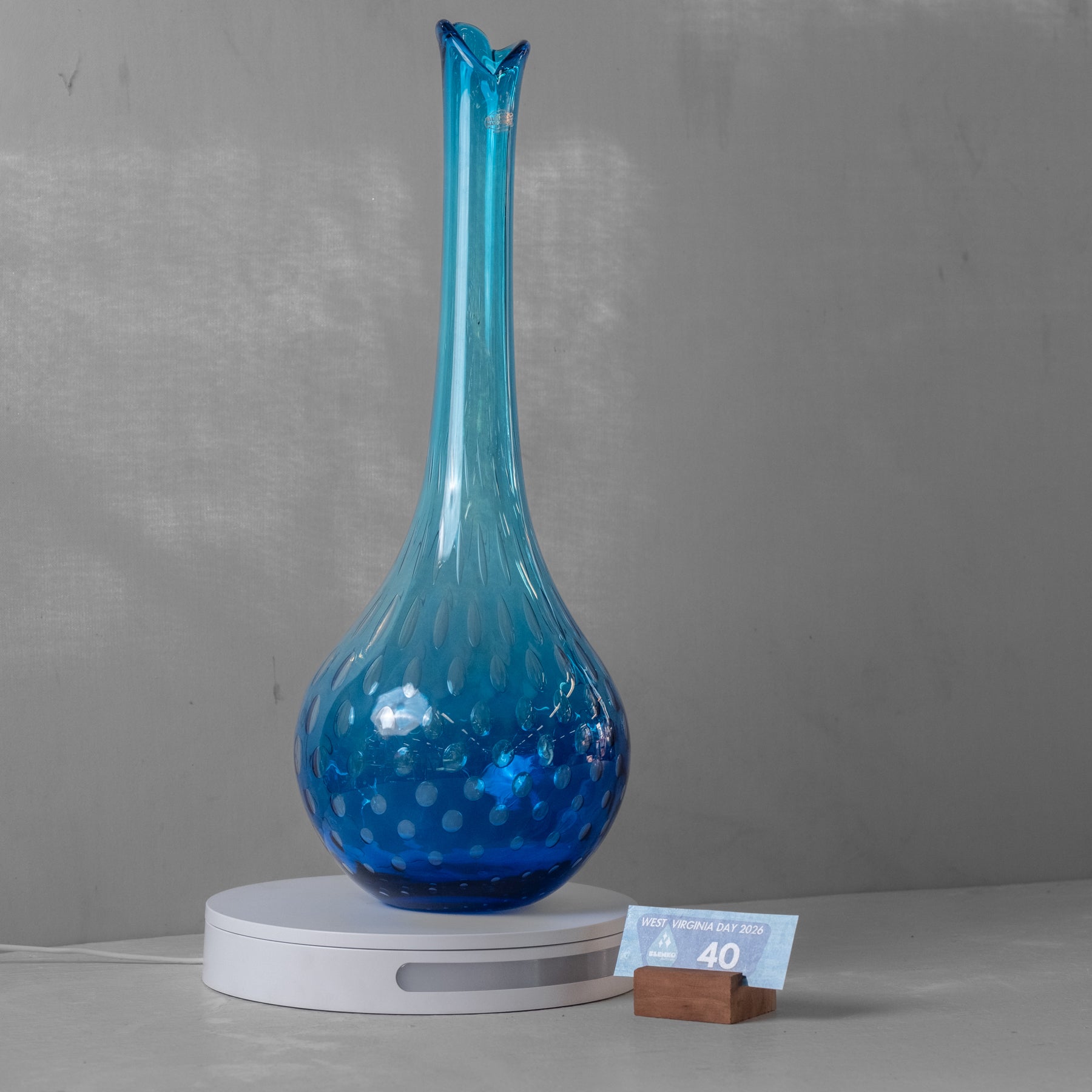 A blue glass vase with a flared neck and bubble pattern, displayed on a white circular light box with a small price card on a wooden stand against a gray wall.