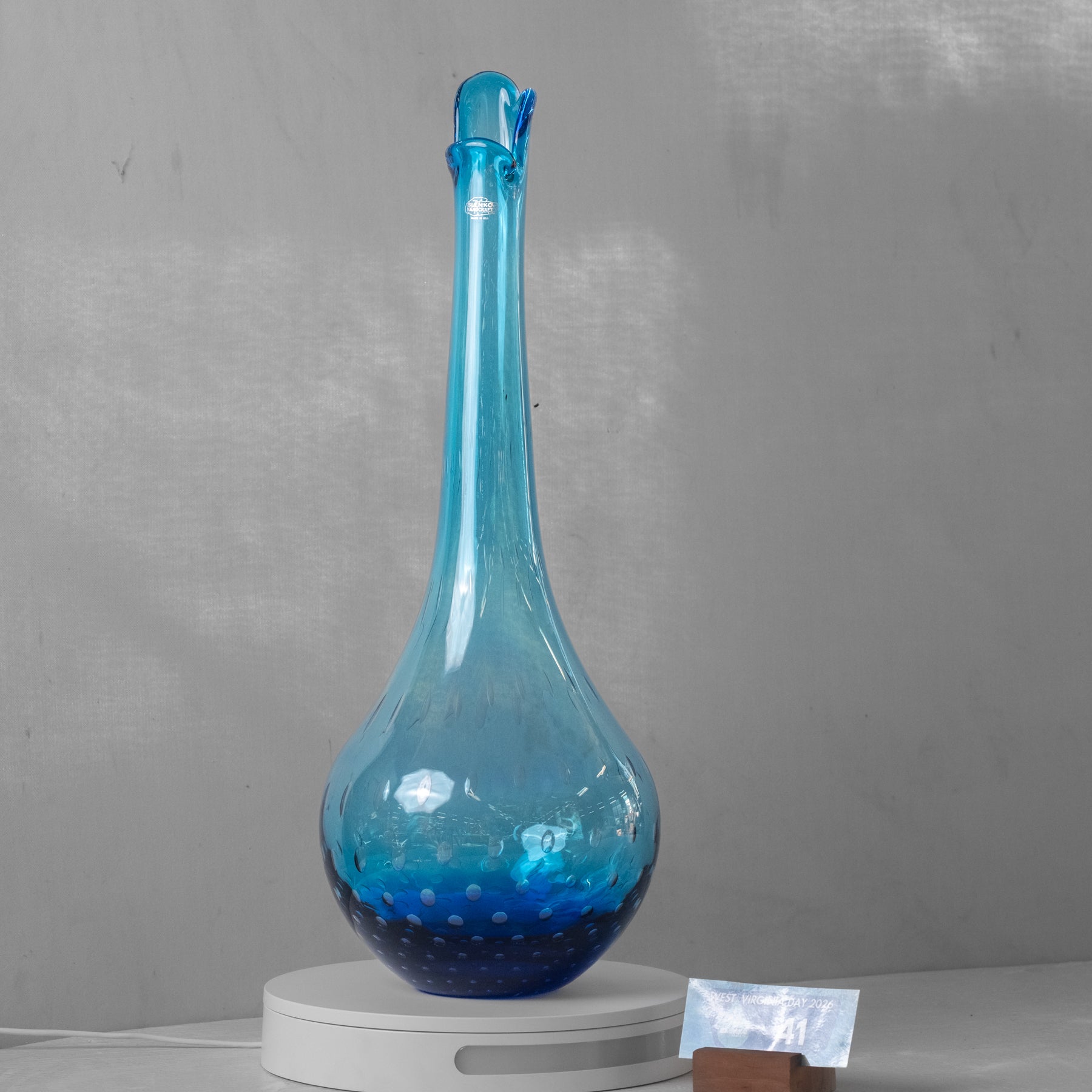 A tall, bulb-shaped blue glass decanter or vase with a narrow neck and small spout, displayed on a white circular base against a gray wall.