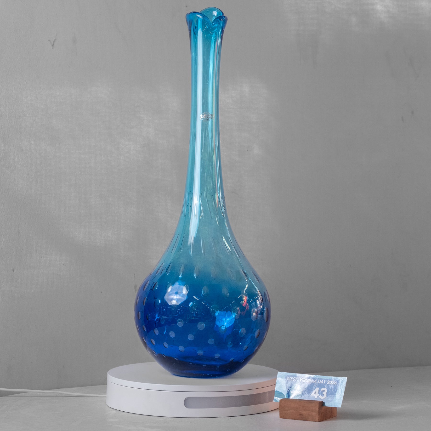 A tall, slender blue glass vase with a flared neck and dotted pattern, displayed on a white circular stand against a gray wall.