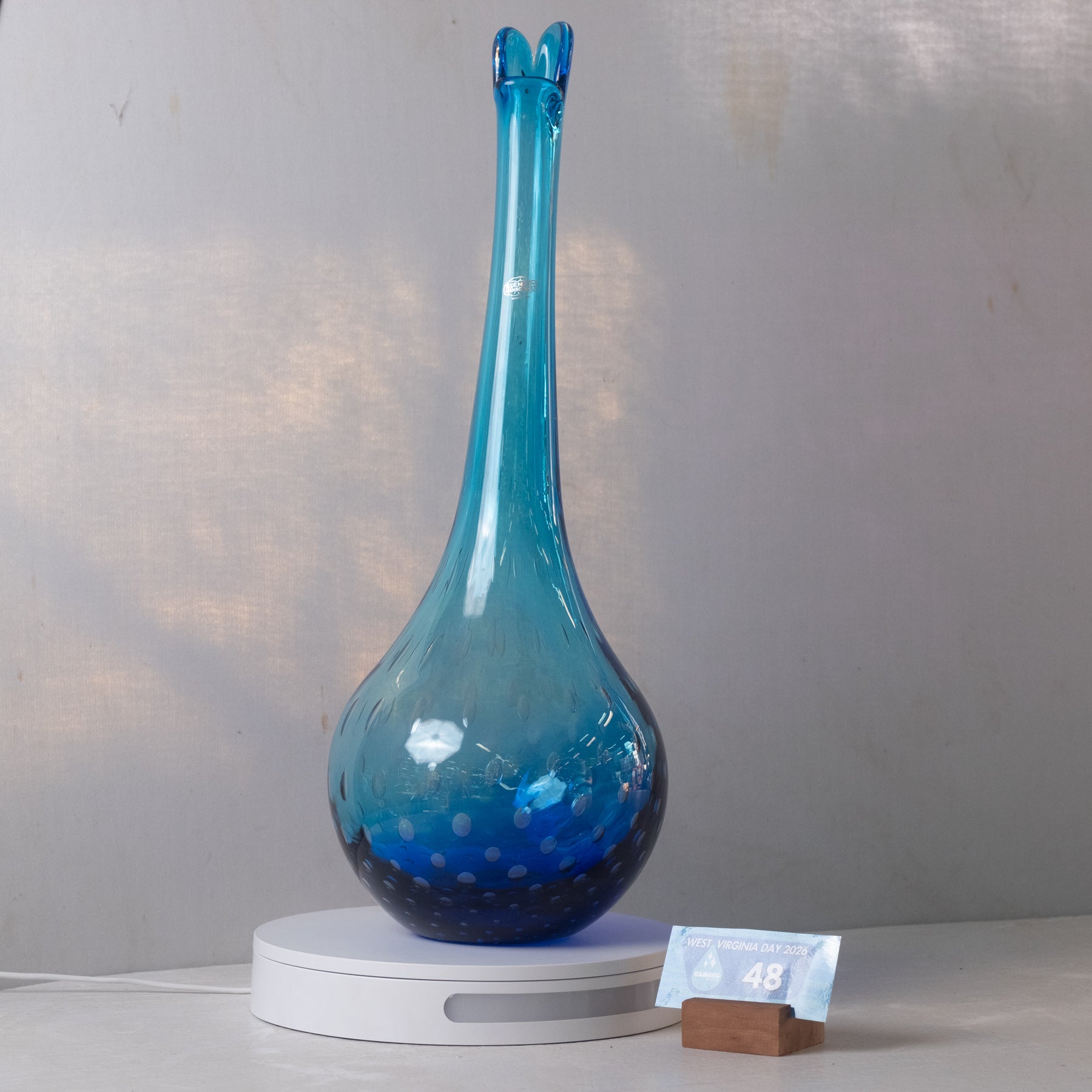 A tall, narrow blue glass vase with a flared rim and subtle bubble pattern, displayed on a white circular light box with a small price tag in front, set against a light gray wall.