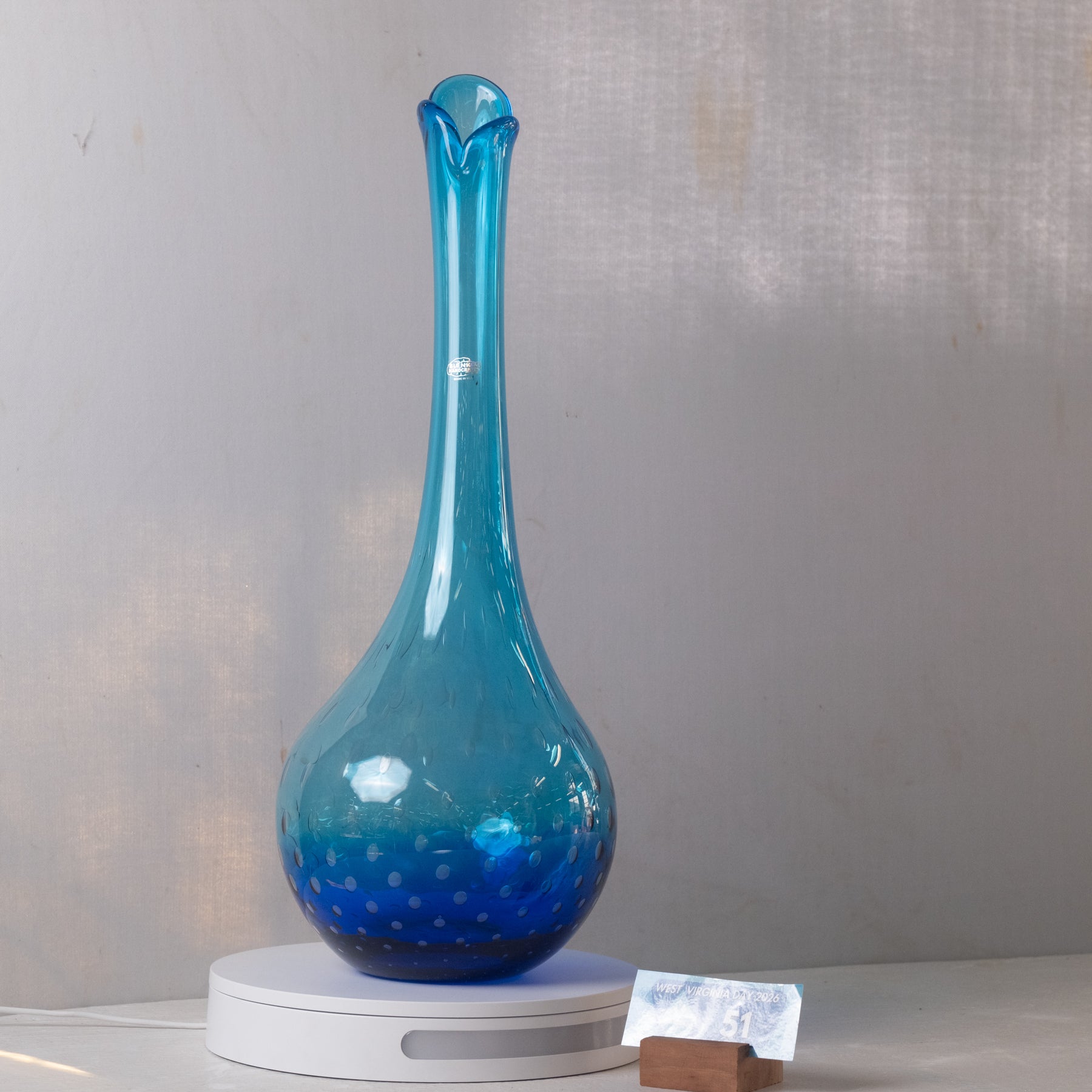 A blue glass decanter with a bulbous base and a narrow flared neck, displayed on a white circular stand against a light gray wall.