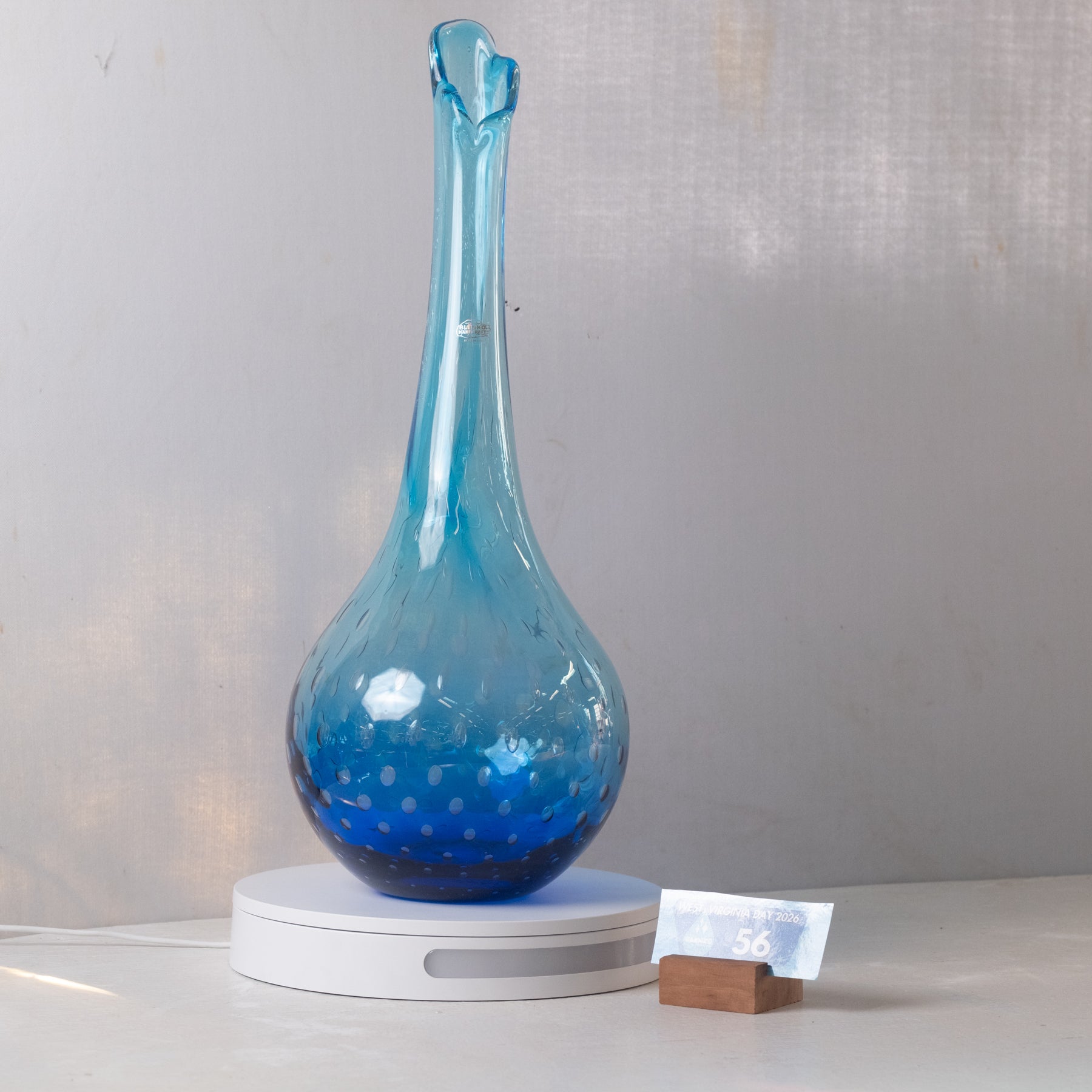 A blue glass decanter with a bulbous body and narrow neck, resting on a white circular base next to a small wooden stand with a card, against a light gray fabric backdrop.