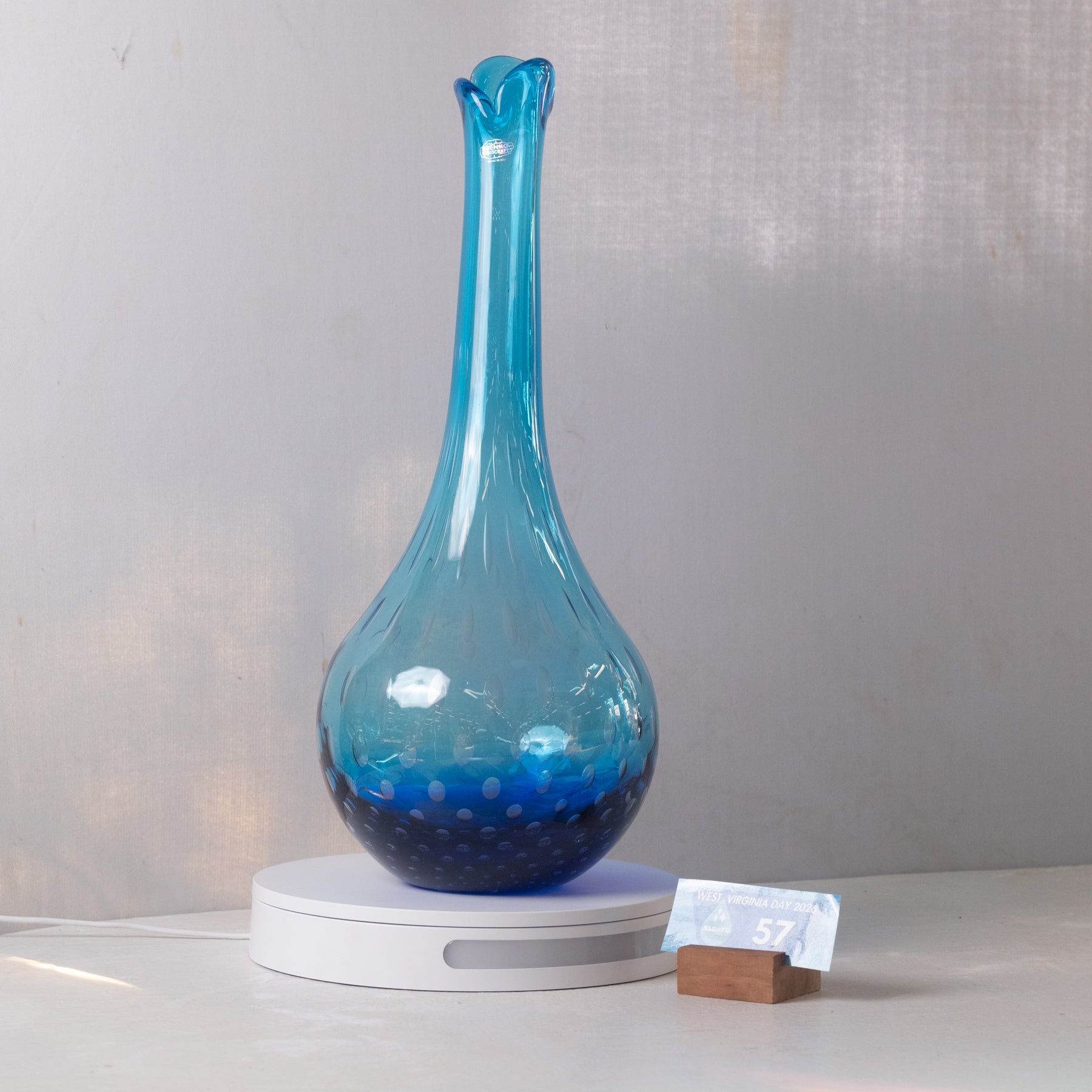 A translucent blue glass vase with a flared neck and dotted pattern concentrated near the base, displayed on a white circular stand against a light gray wall.