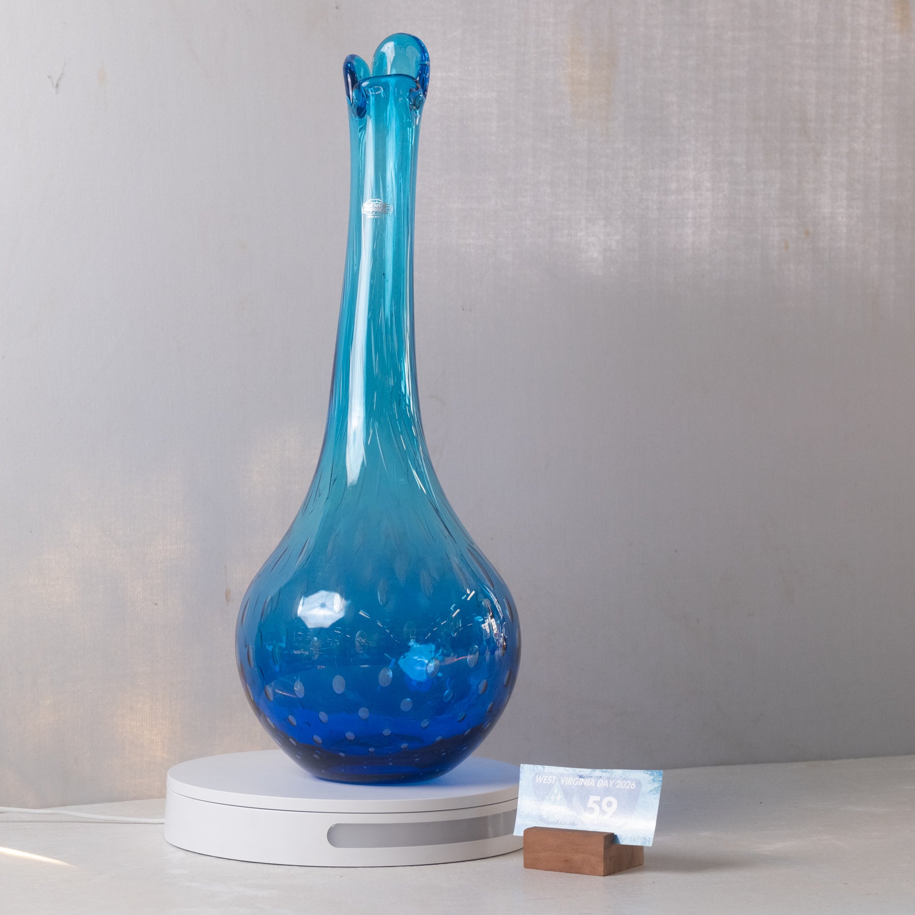A translucent blue glass vase with a long, flared neck and bubble-like rim, displayed on a white circular lighted base against a light gray wall.
