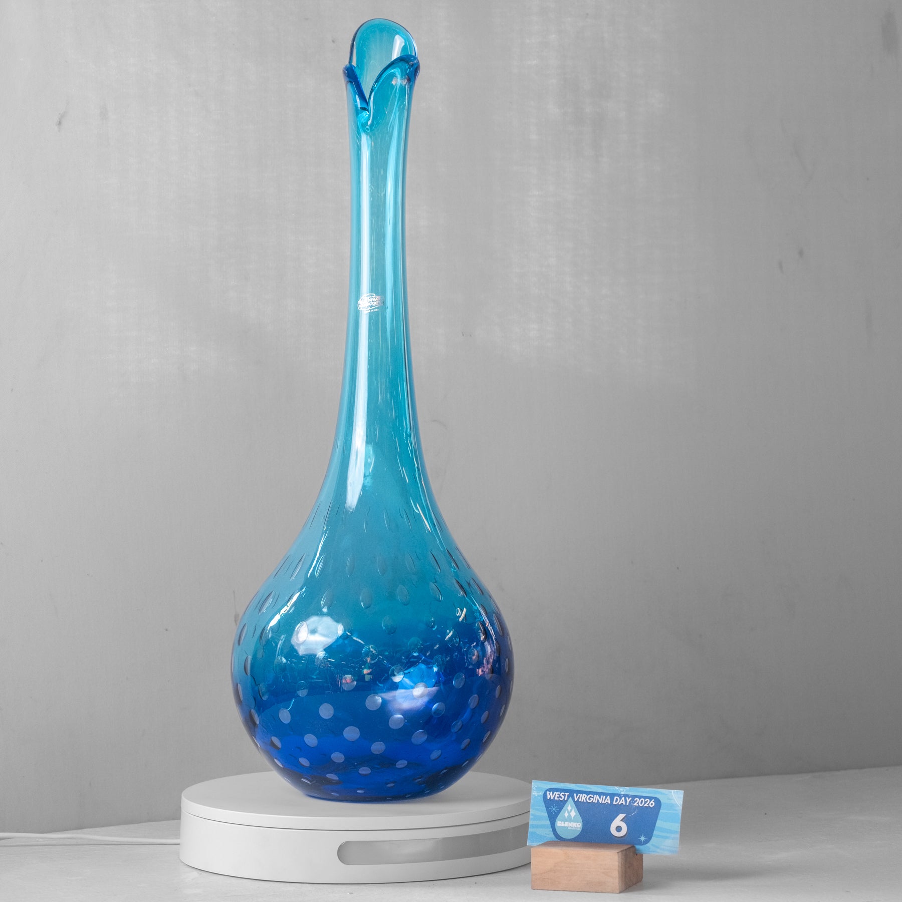 A blue glass decanter with a long neck and dotted pattern, displayed on a white circular charging base next to a small card, against a gray wall.