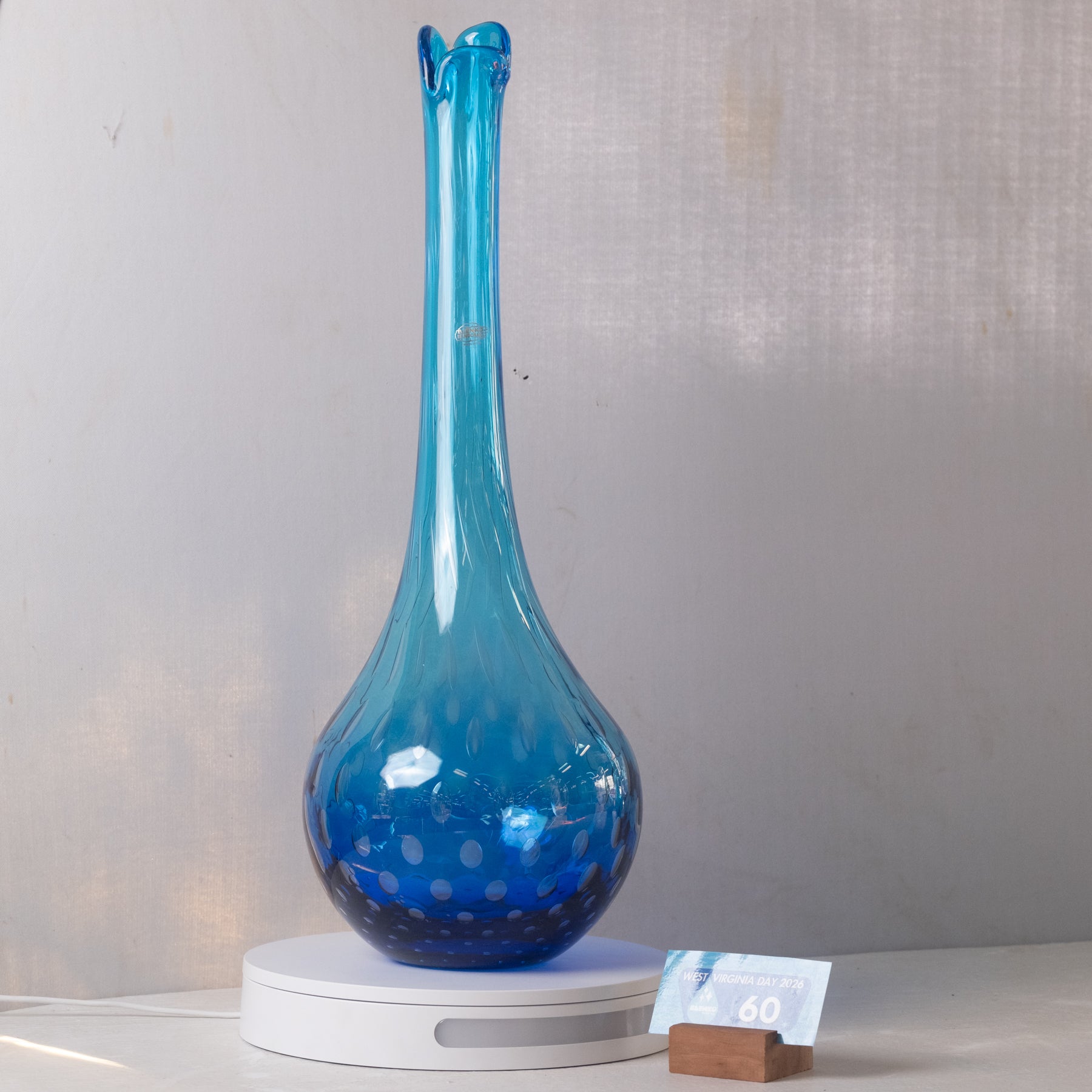 A tall, bulb-shaped blue glass vase with a flared neck and spotted pattern, displayed on a white circular stand against a light gray wall.